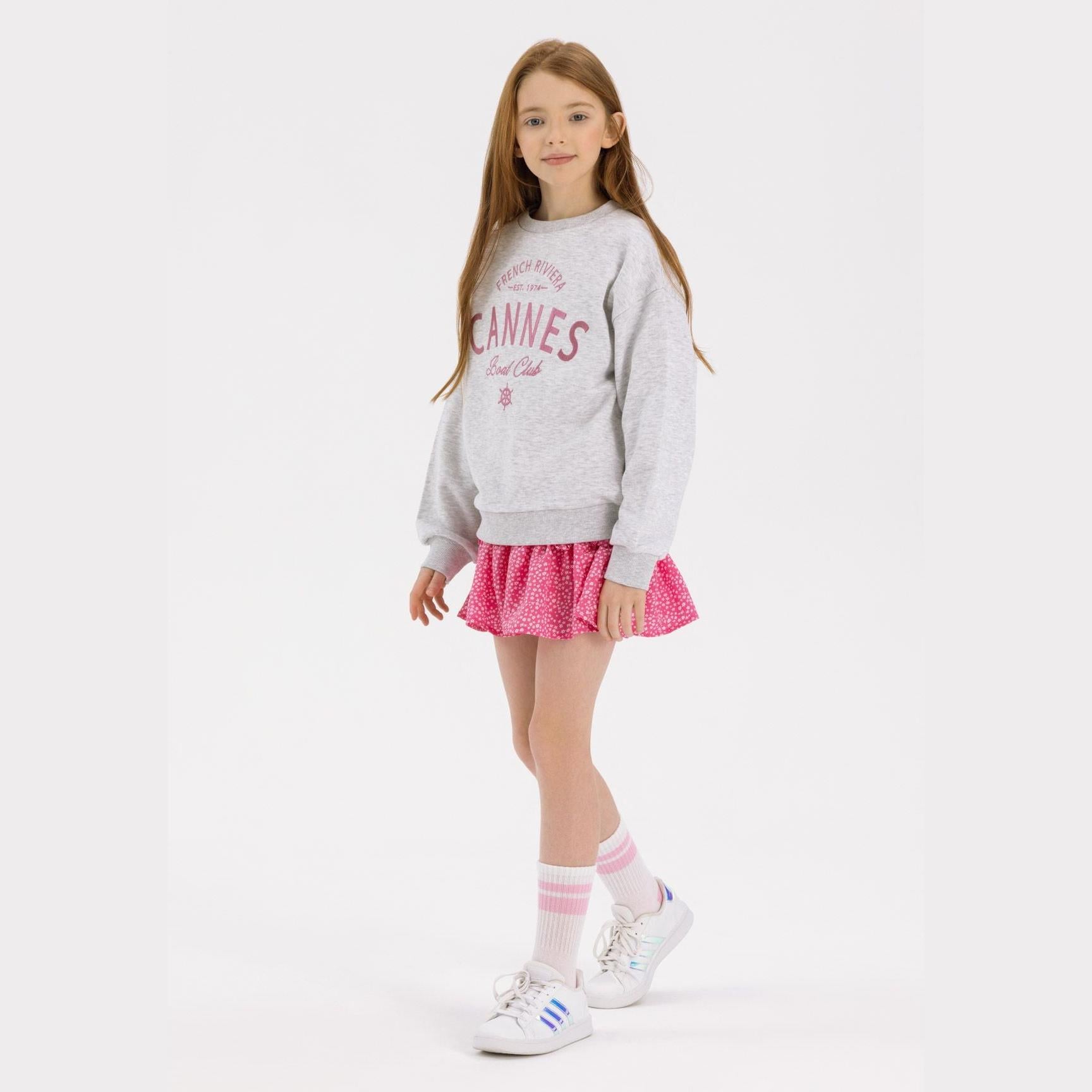 Young girl wearing grey fleece sweatshirt with pink Cannes Boat Club print, paired with pink polka dot skirt and white sneakers on white background