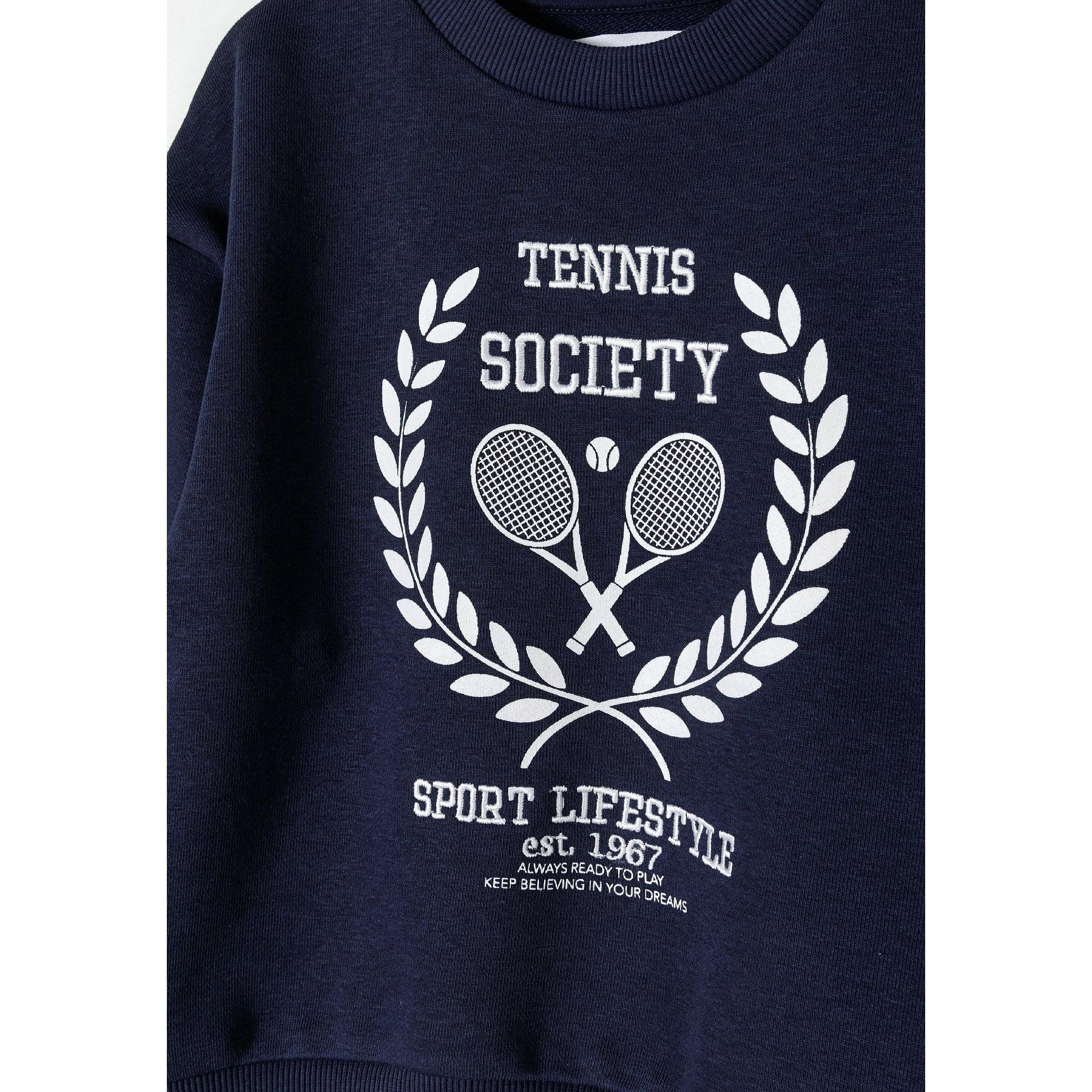 Close-up detail of tennis society print on navy sweatshirt showing crossed rackets, tennis ball and laurel wreath with text