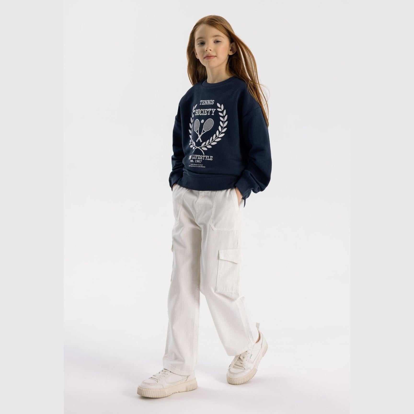 Girl wearing navy fleece sweatshirt with tennis society print and white cargo trousers, standing against white background