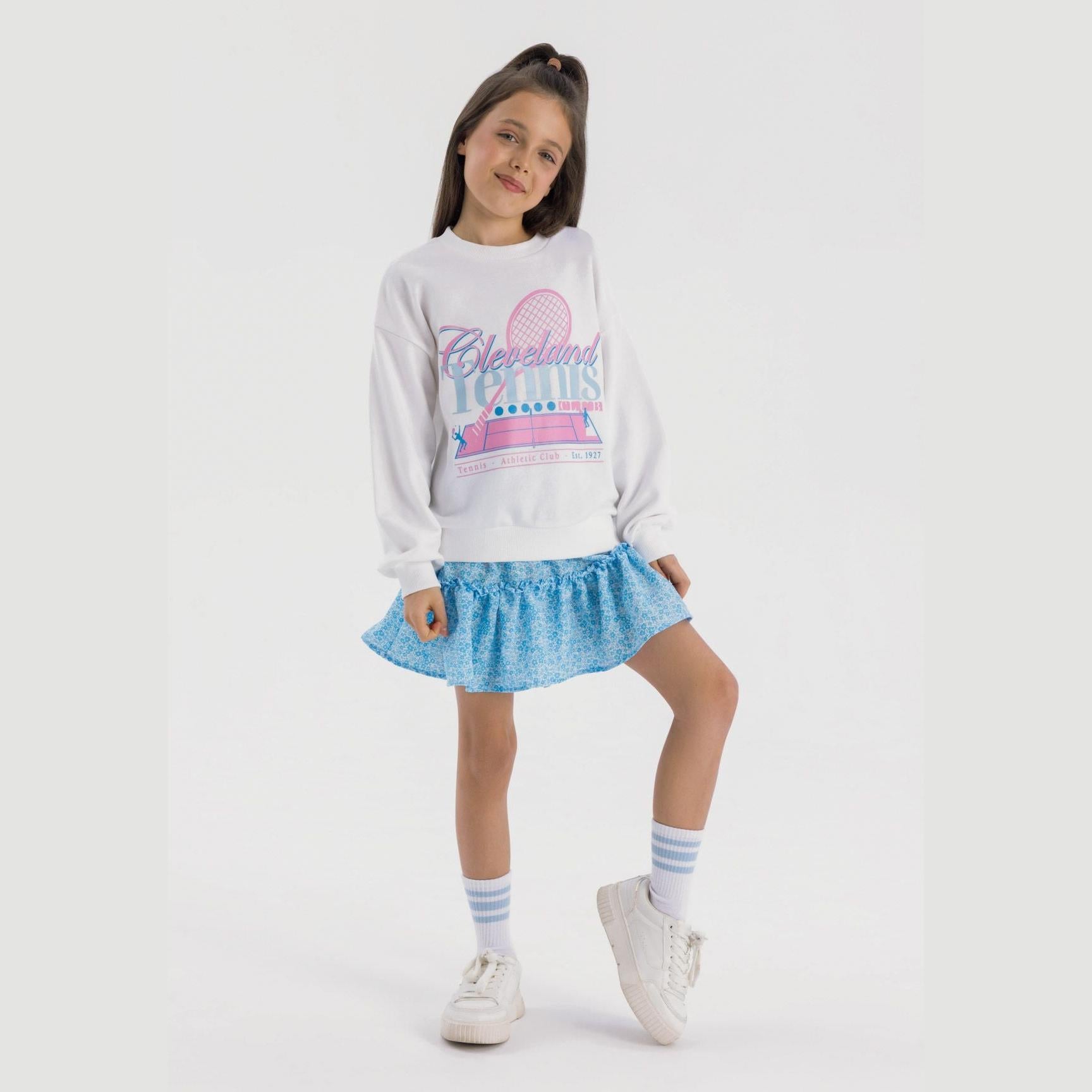 Girl wearing cream fleece sweatshirt with Cleveland Tennis print, paired with blue patterned skirt and striped socks against white background