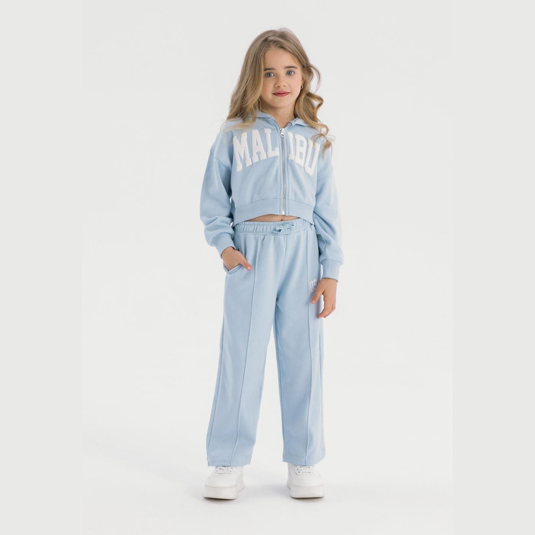 Young girl wearing light blue tracksuit set with cropped zip hoodie and wide leg joggers, standing against white background with white trainers