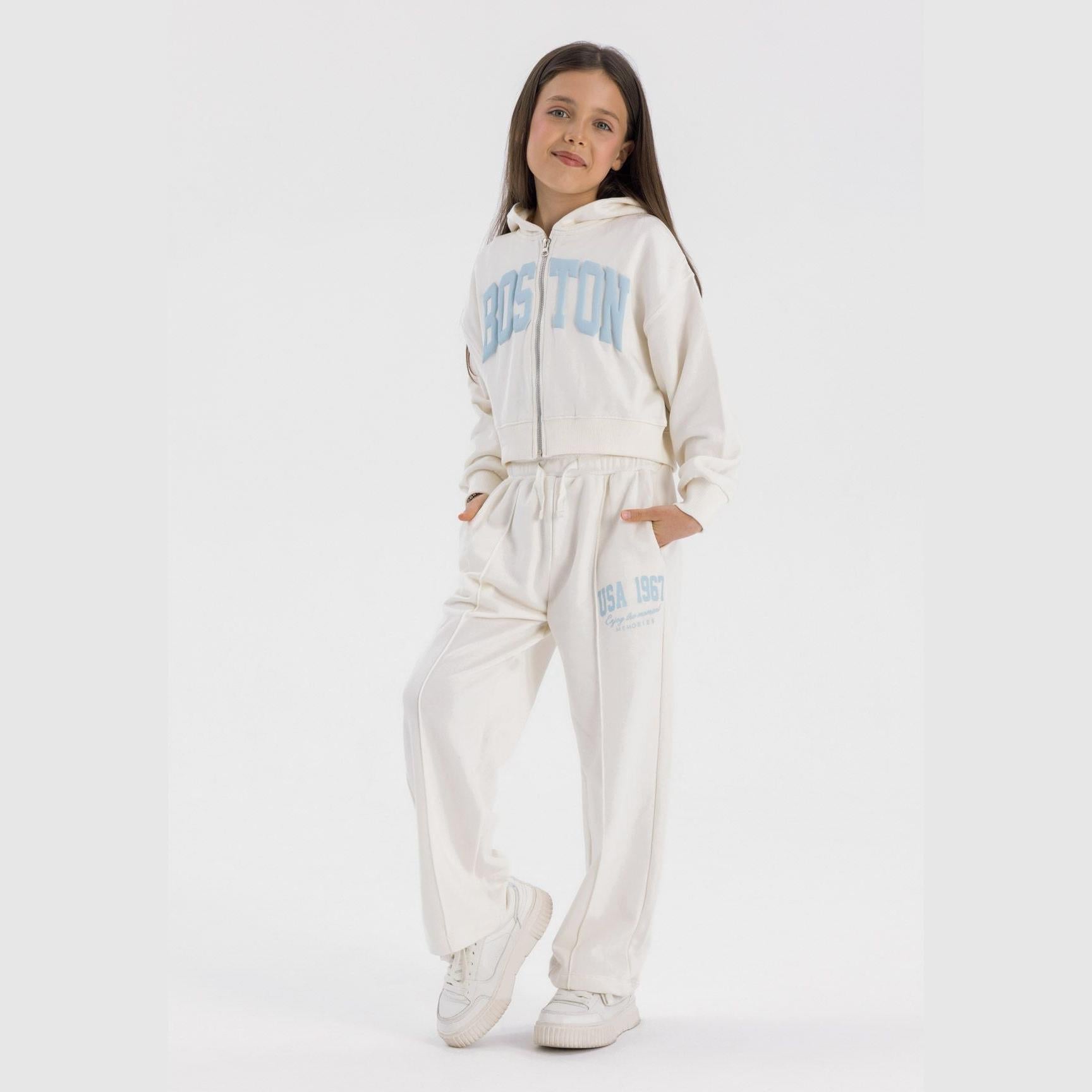 Girl wearing cream tracksuit set with cropped hoodie featuring blue text print and wide leg joggers, standing against light grey background