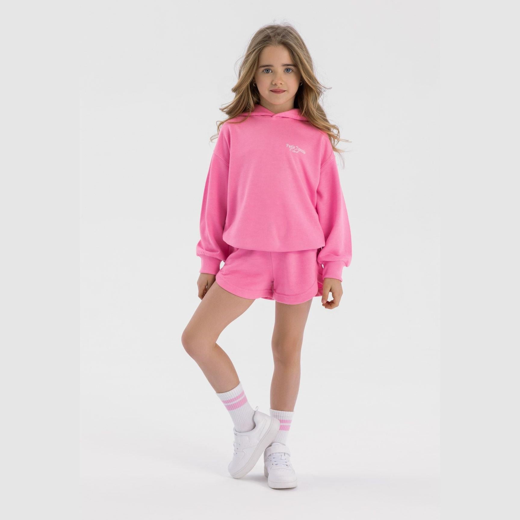 Young girl wearing pink hoodie and shorts set, standing against white background, styled with white sneakers and pink striped socks