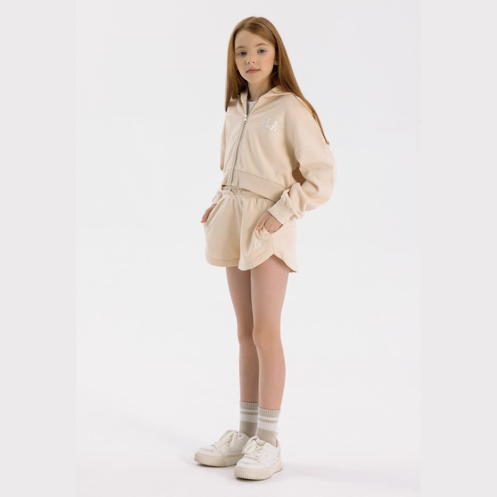 Young girl wearing beige zip hoodie and shorts set, standing against white background, styled with white sneakers and striped socks