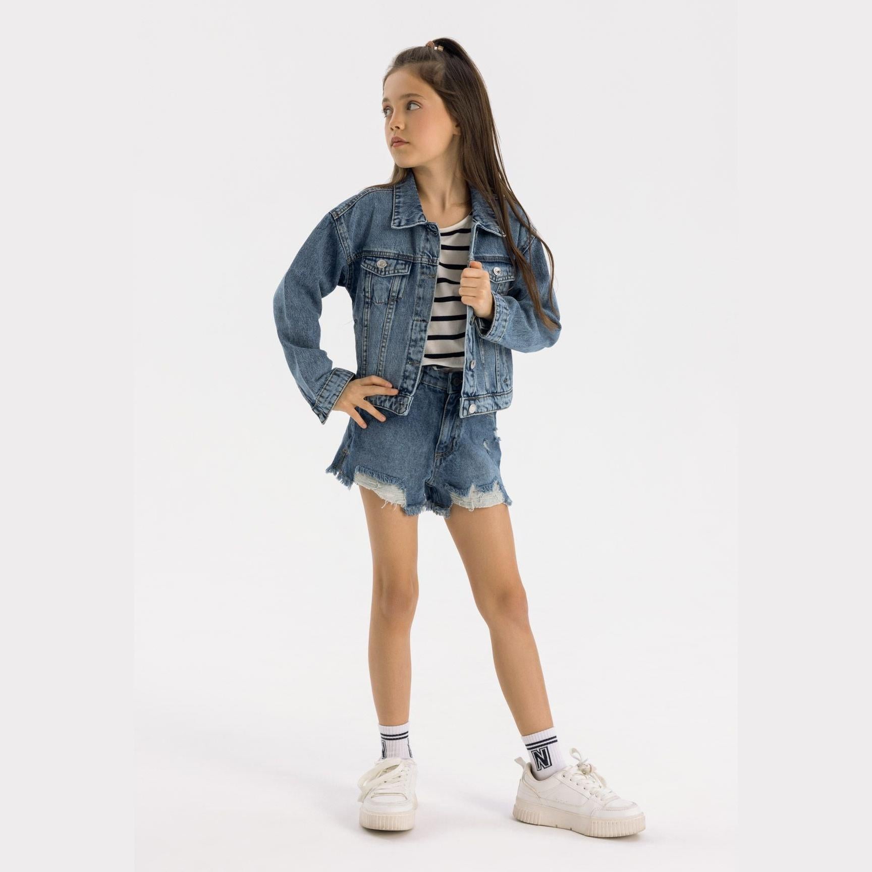 Young girl wearing oversized navy denim jacket over striped top with denim shorts, white sneakers, standing against white background
