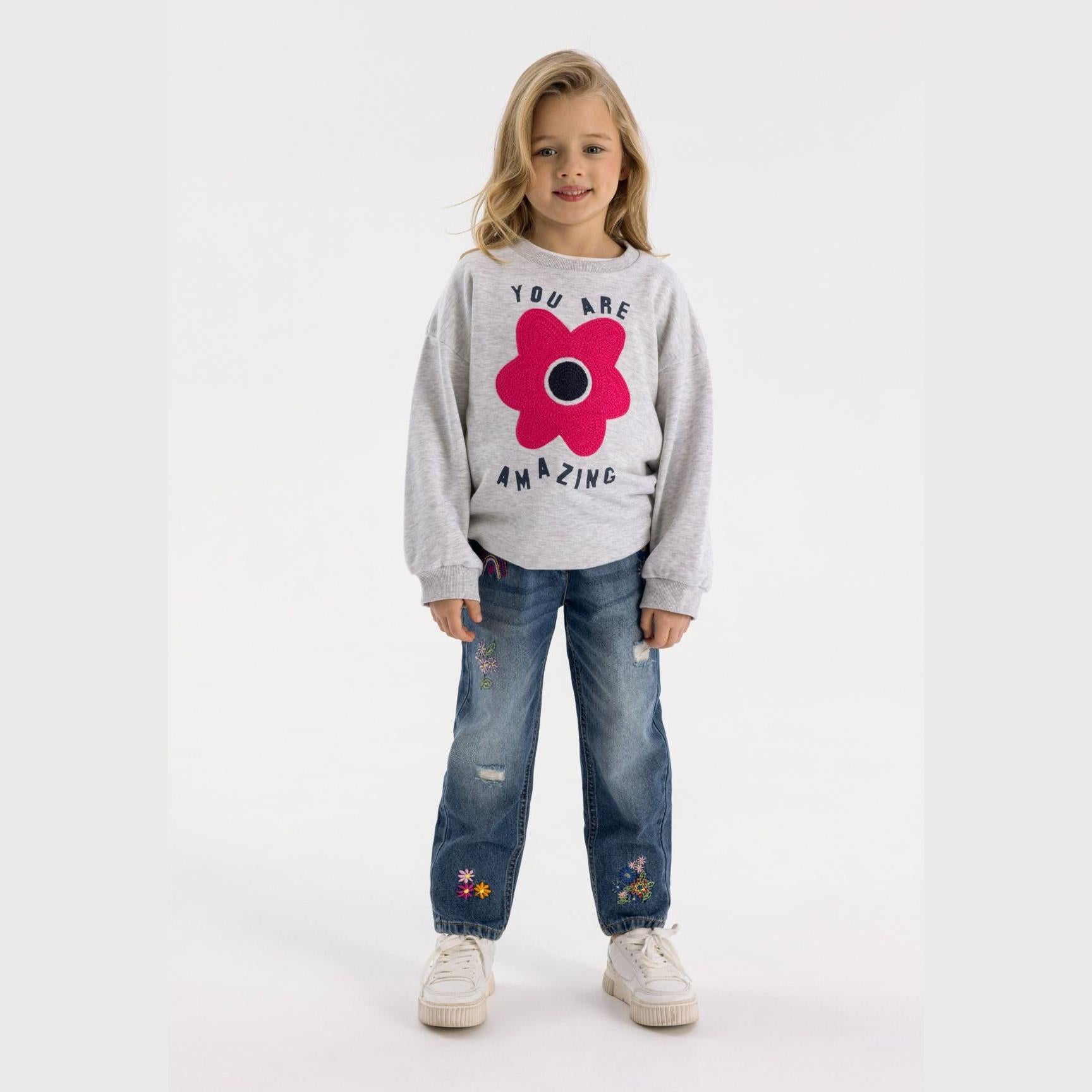 Young girl wearing grey sweatshirt with pink flower design and navy embroidered jeans with white sneakers on light background