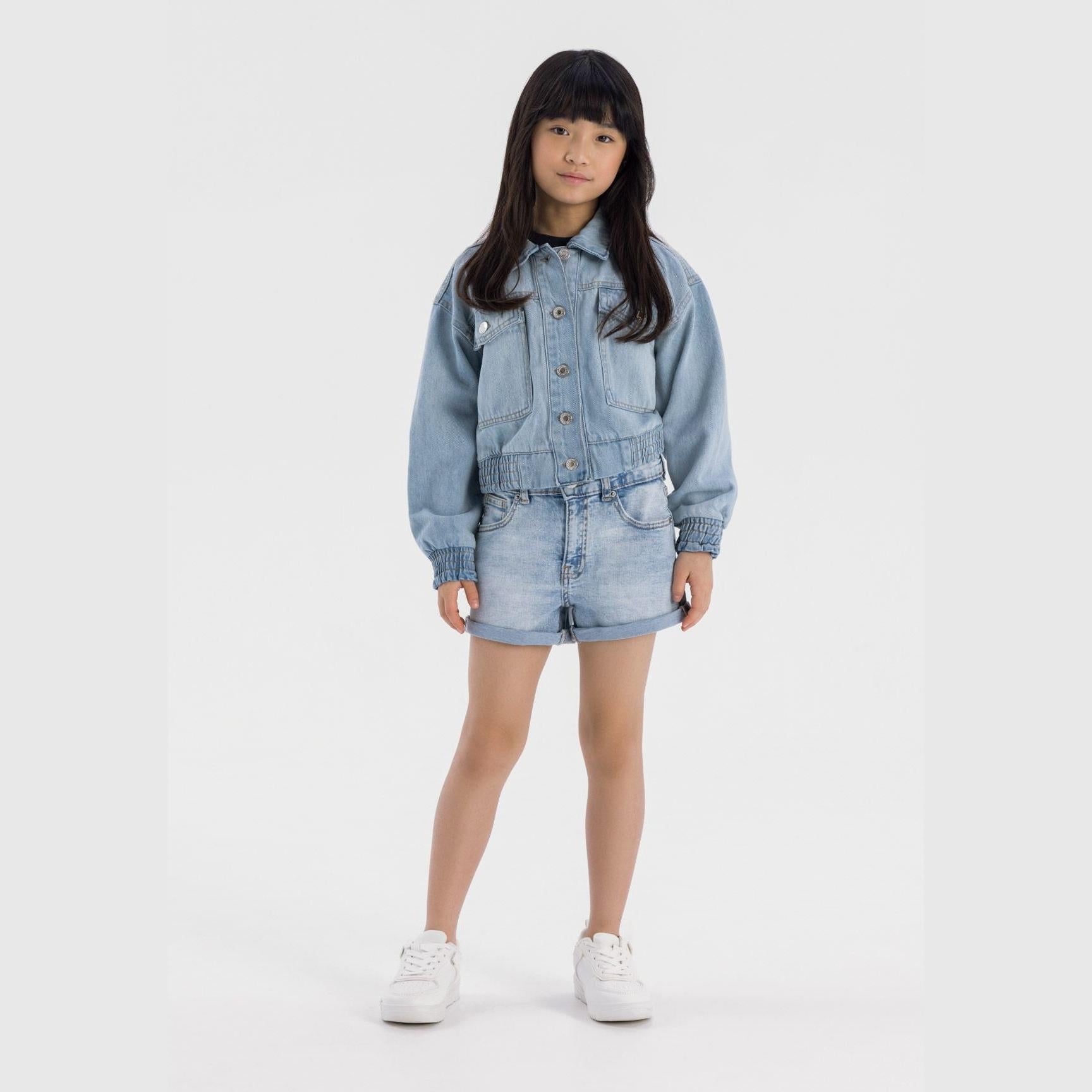 Young girl wearing light blue high-waisted denim shorts with matching denim jacket, standing against white background