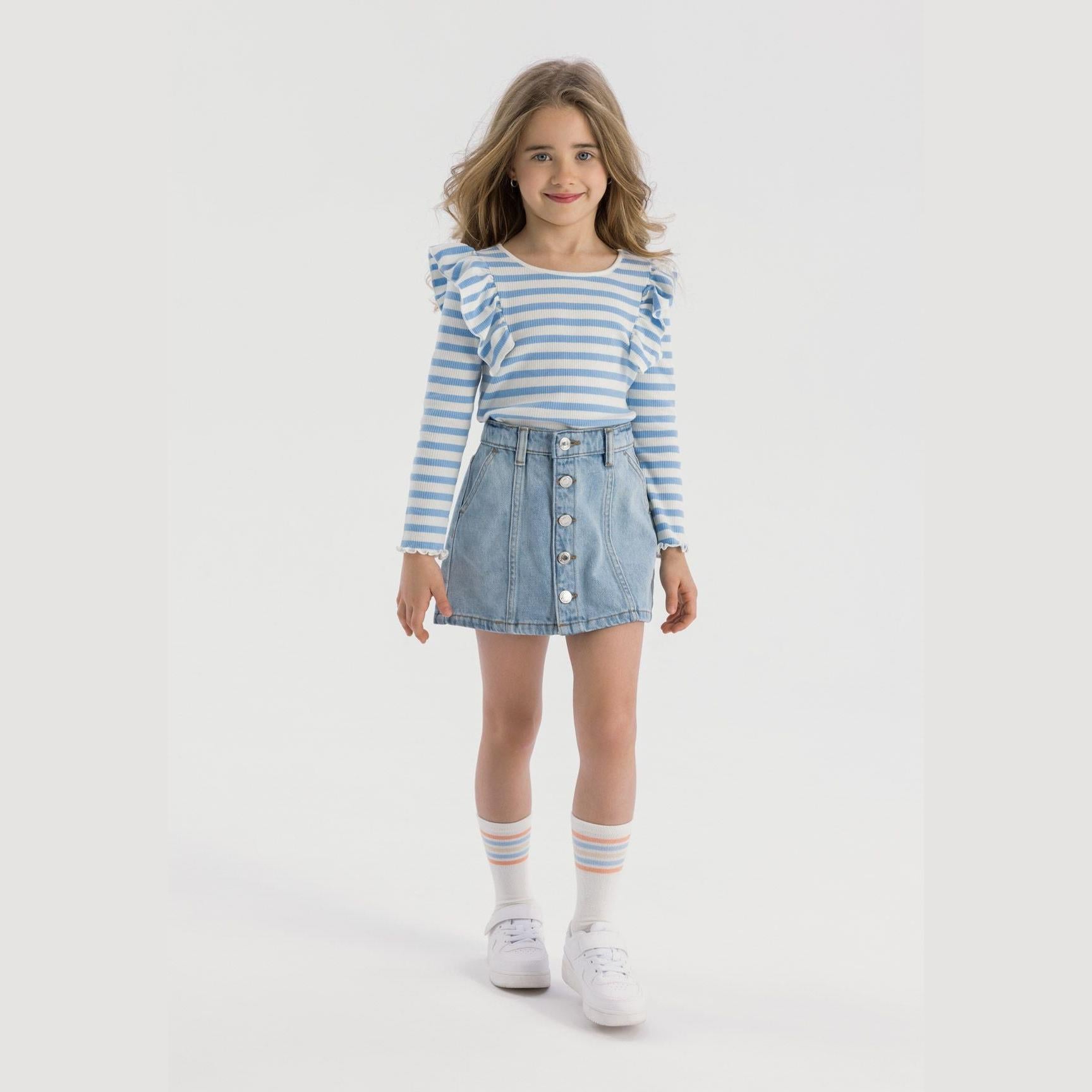 Young girl wearing light blue denim skirt with button fastening and blue striped ruffle sleeve top, white socks with blue stripes and white sneakers on white background