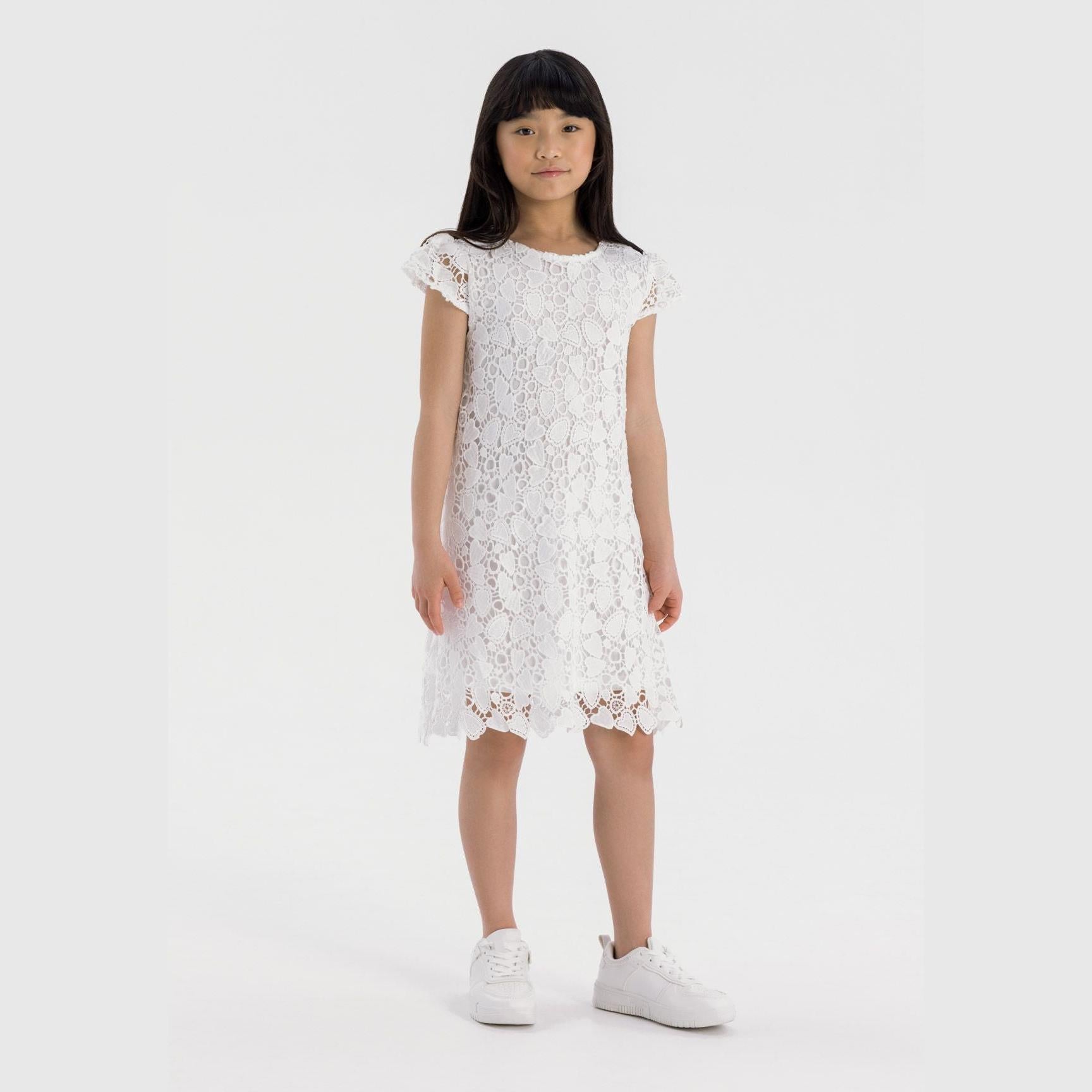 Young girl wearing white lace dress with intricate floral patterns, short sleeves, and knee-length hem, standing against light grey background wearing white sneakers