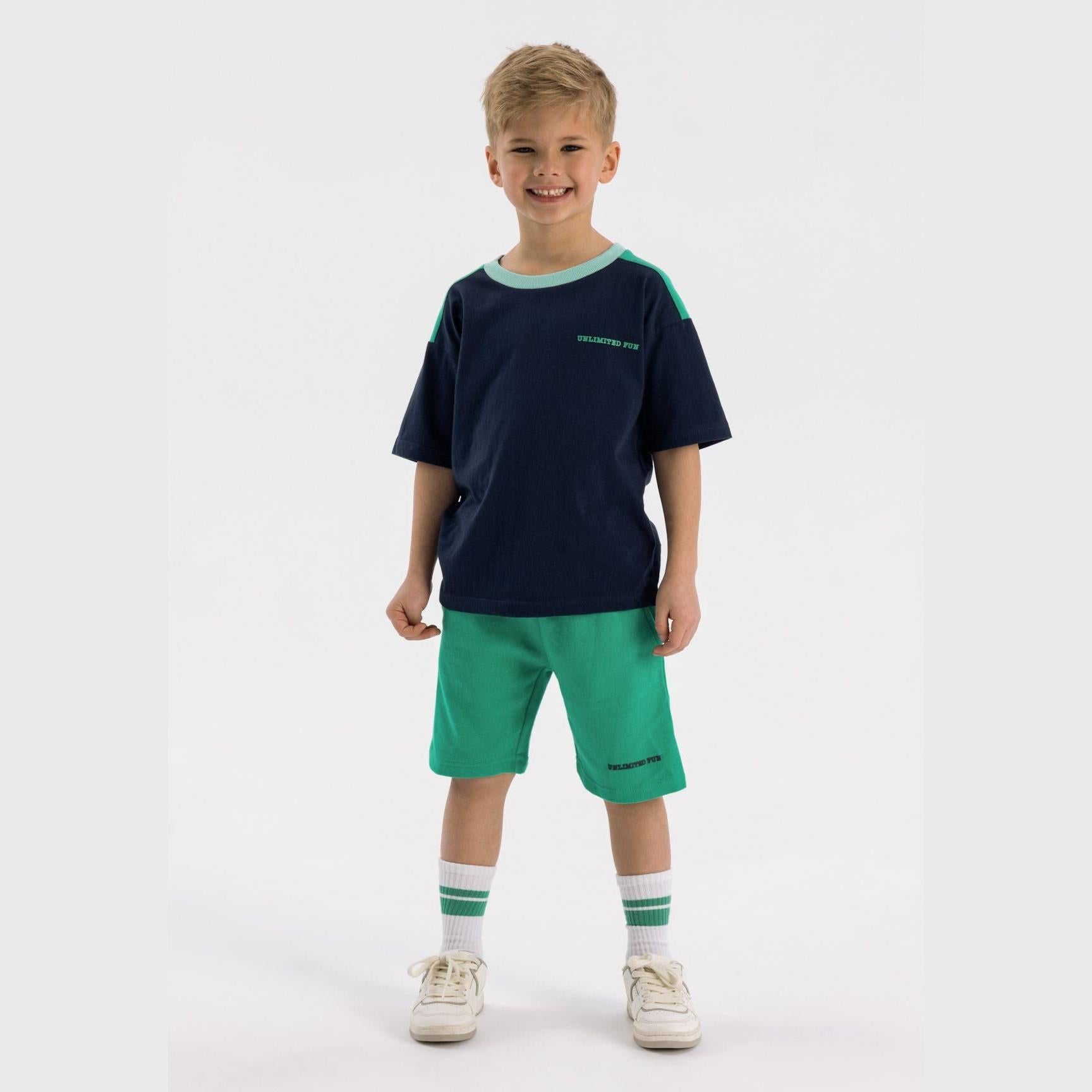 Young boy wearing navy t-shirt with mint green trim and bright green fleece shorts, standing against white background with striped socks and trainers