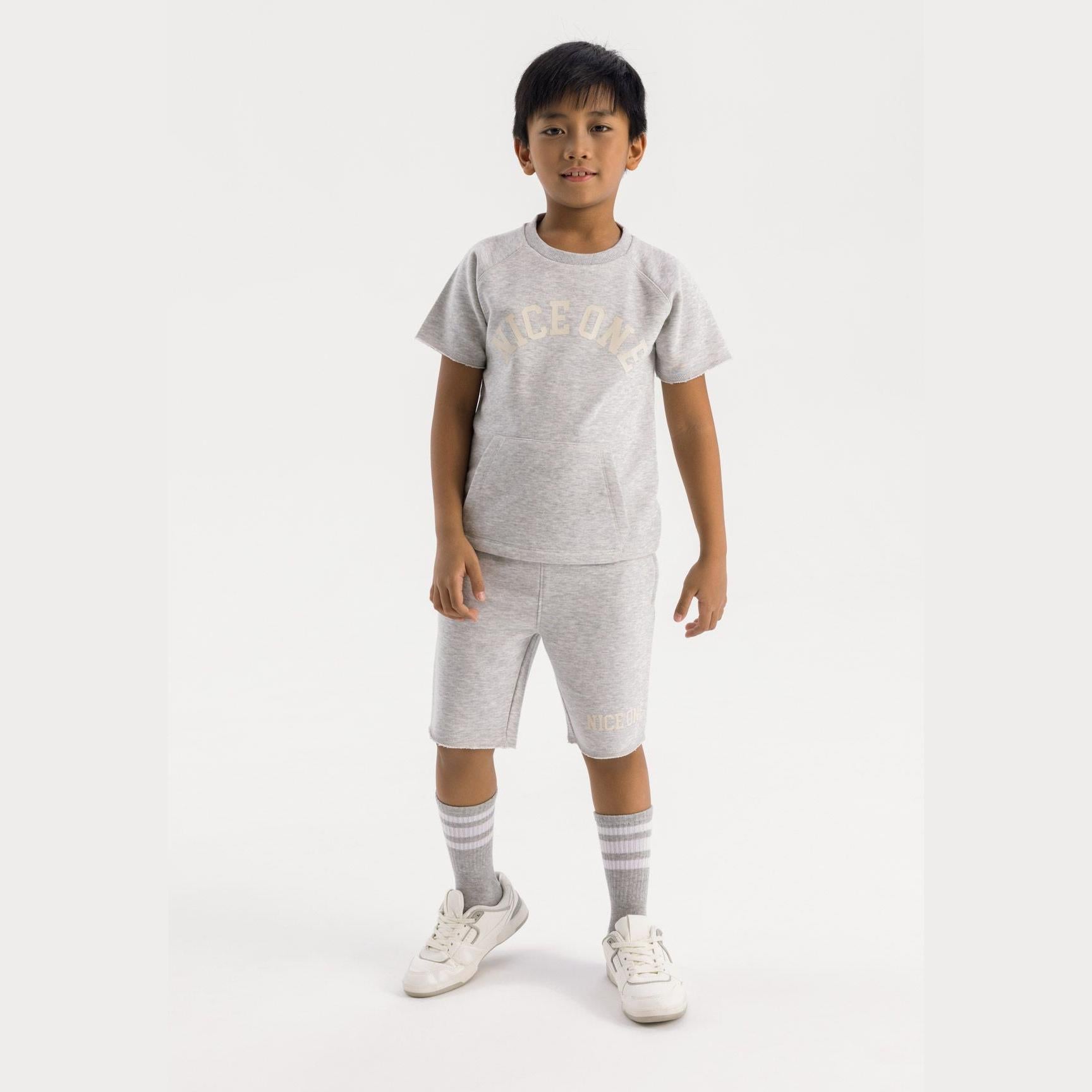 Young boy wearing grey jersey t-shirt and shorts set with Nice One print, standing against white background with grey striped socks and white trainers