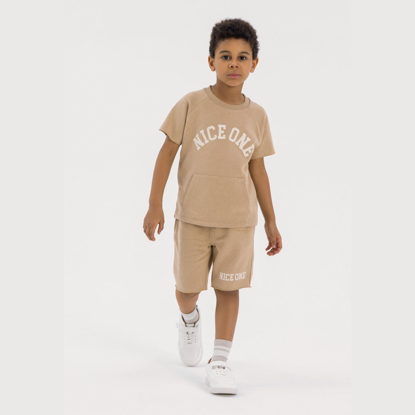 Young boy wearing beige fleece t-shirt and shorts set with Nice One print, standing against white background wearing white trainers