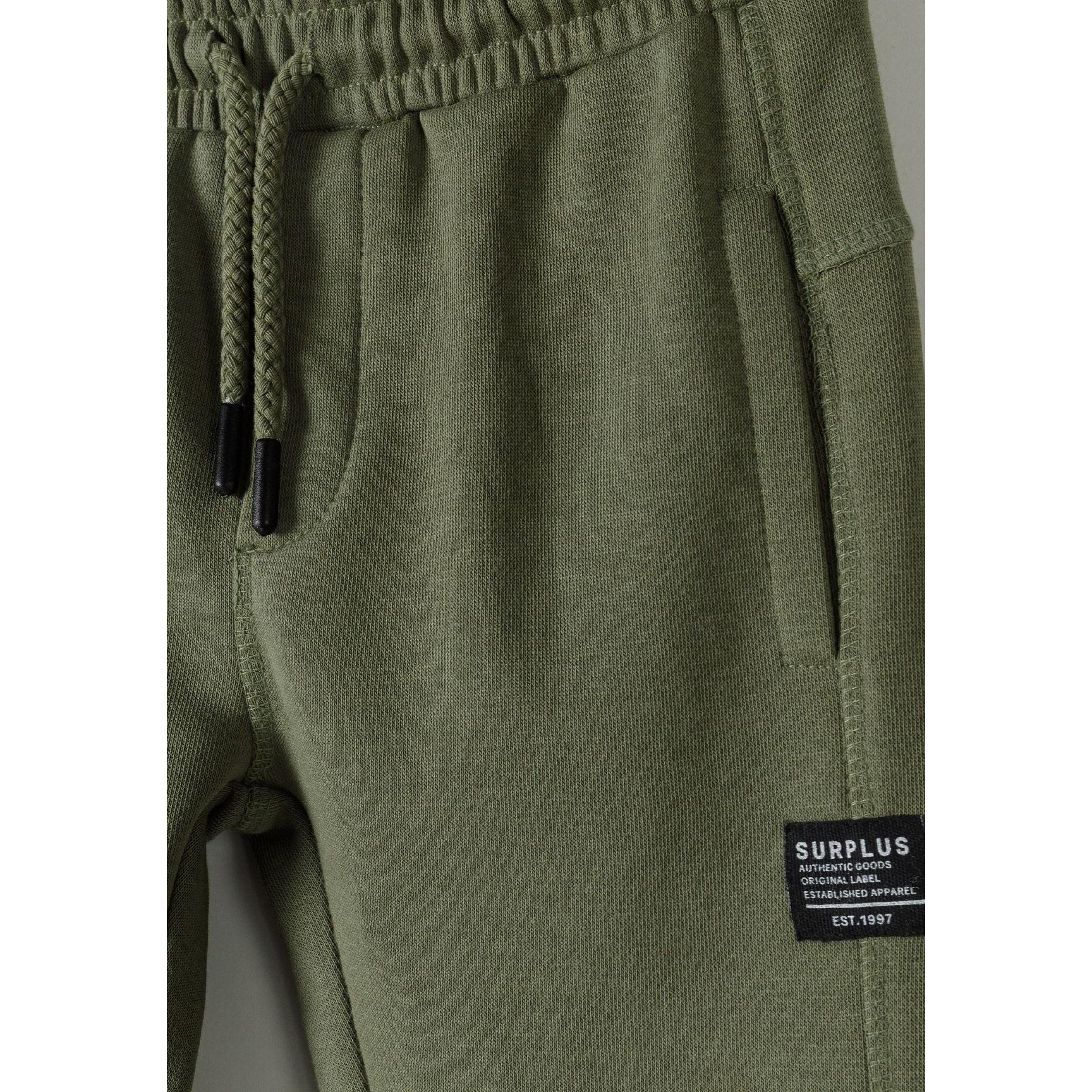 Close-up detail of khaki sweatpants showing side pocket with SURPLUS label and drawstring waistband