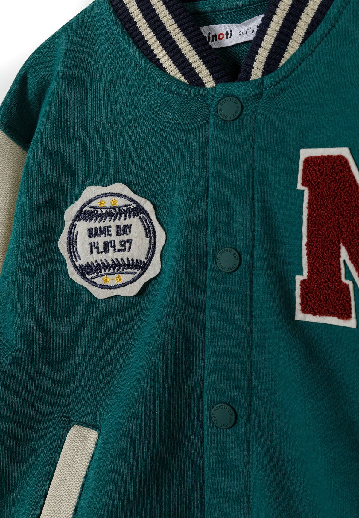 Close-up detail of green baseball jacket showing snap buttons, 'M' logo patch and game day badge