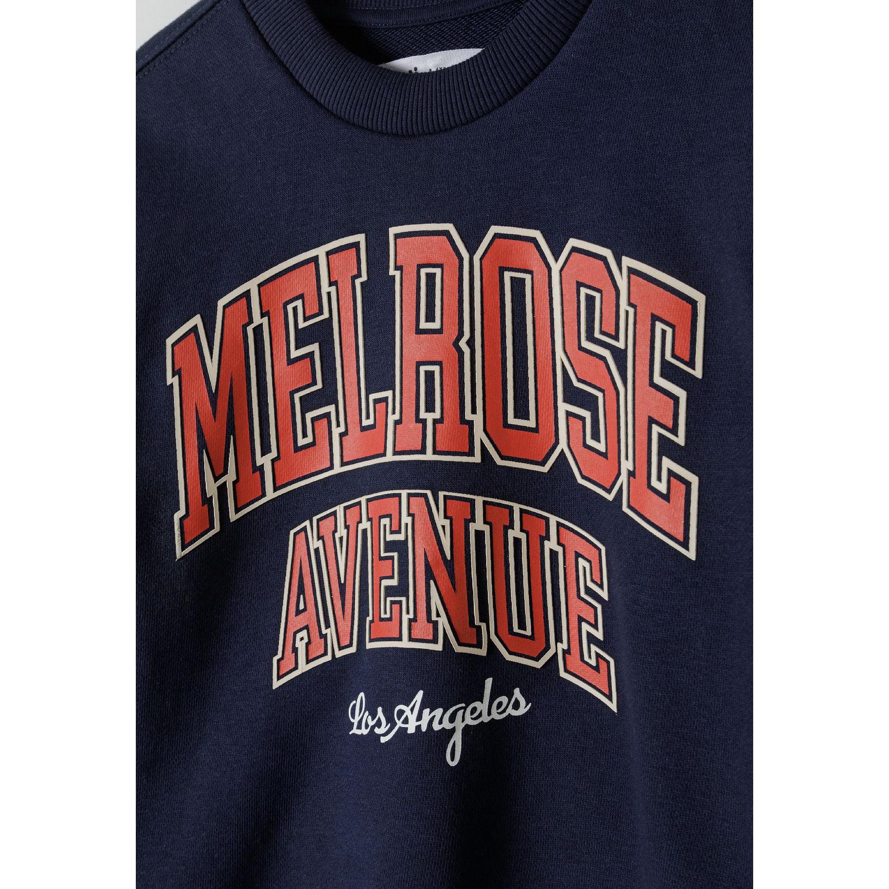 Close-up detail of navy sweatshirt showing Melrose Avenue print in red and white letters with Los Angeles script underneath, crew neck collar visible