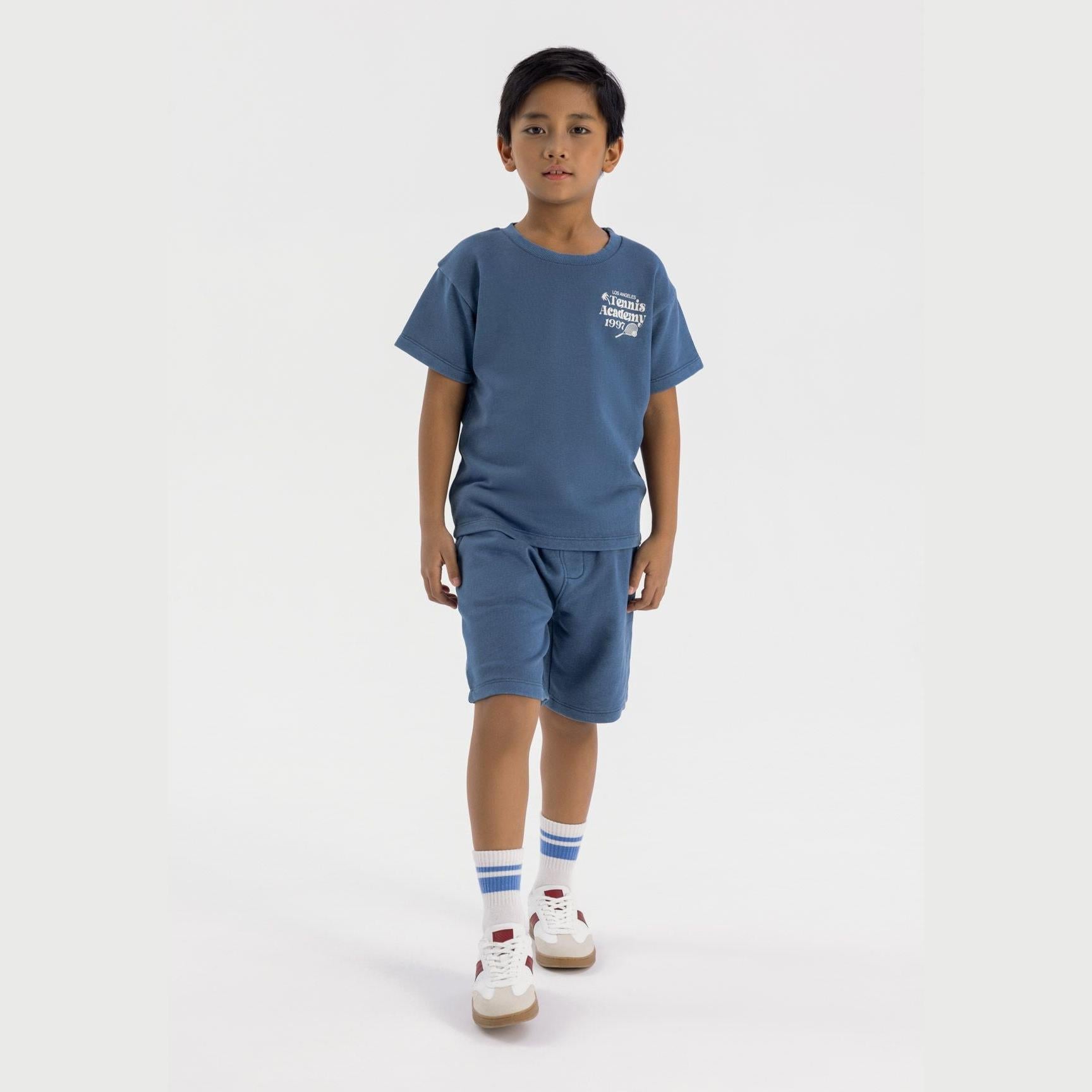 Boy wearing blue tennis academy t-shirt and matching shorts set, standing against white background with white sneakers and striped socks