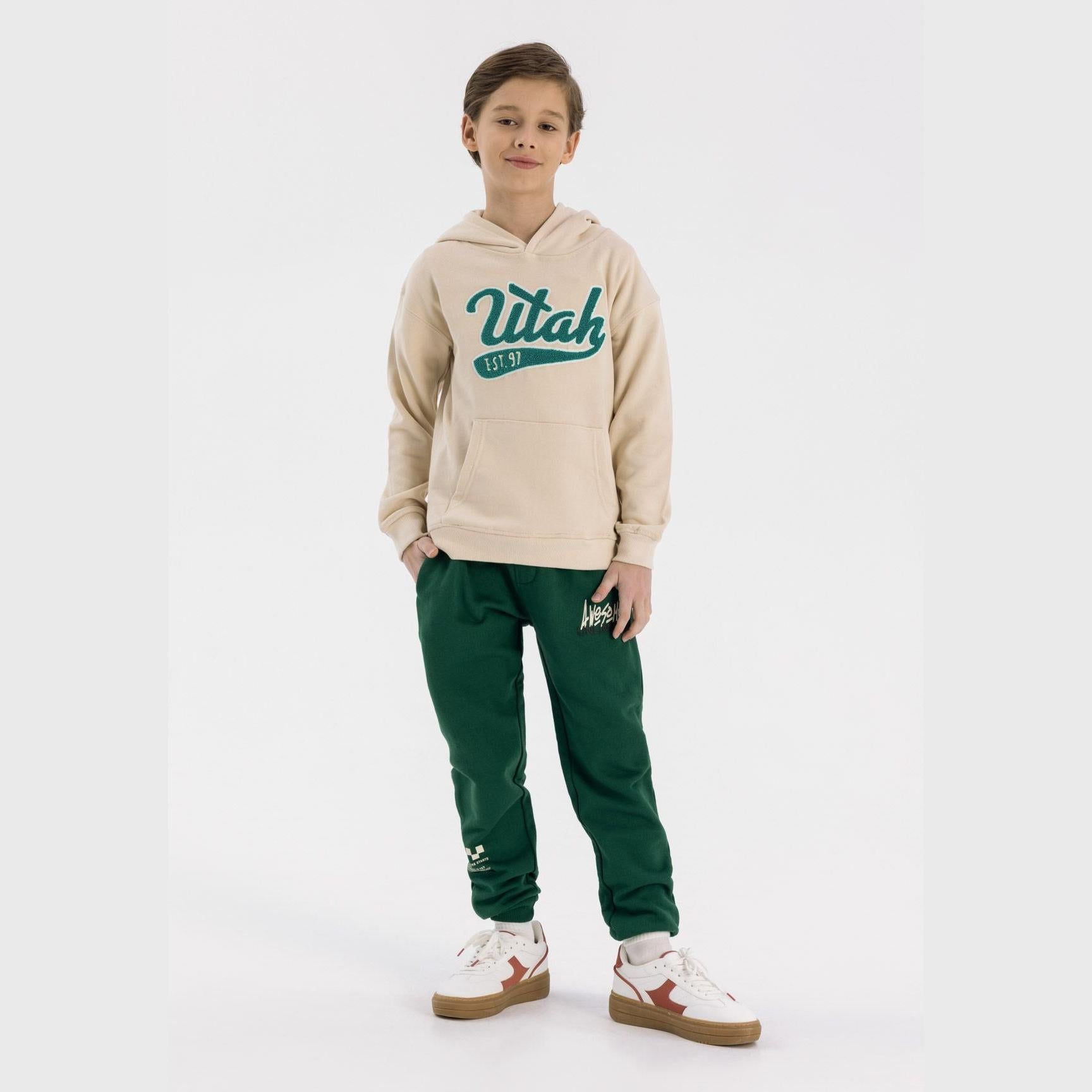 Boy wearing cream oversized hoodie with Utah logo and green joggers against white background