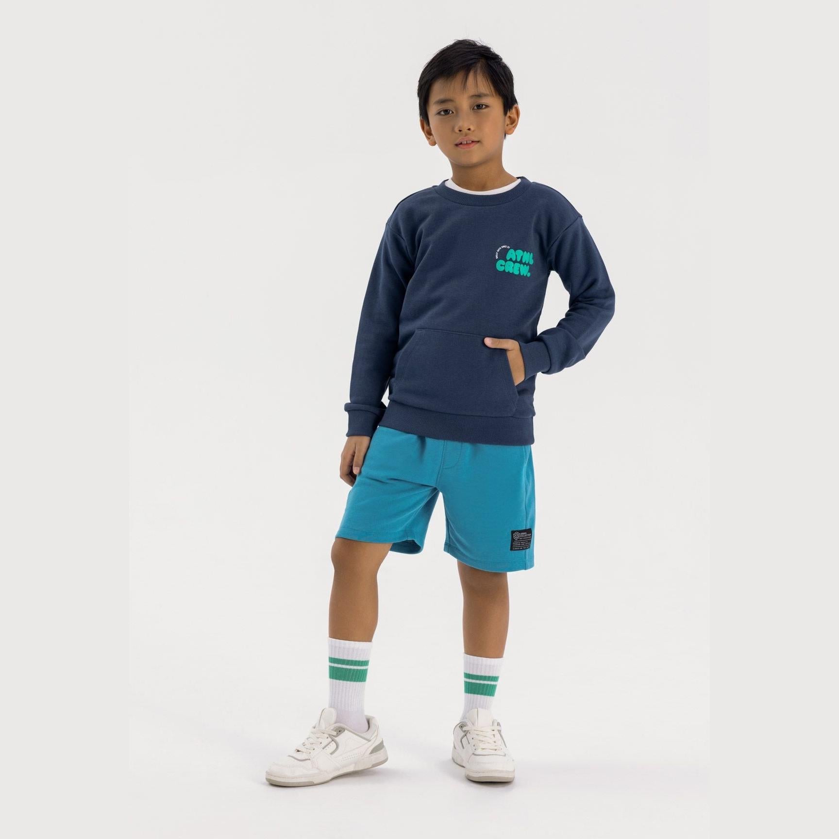 Boy wearing blue fleece shorts with navy sweatshirt, standing against white background showing full outfit styling