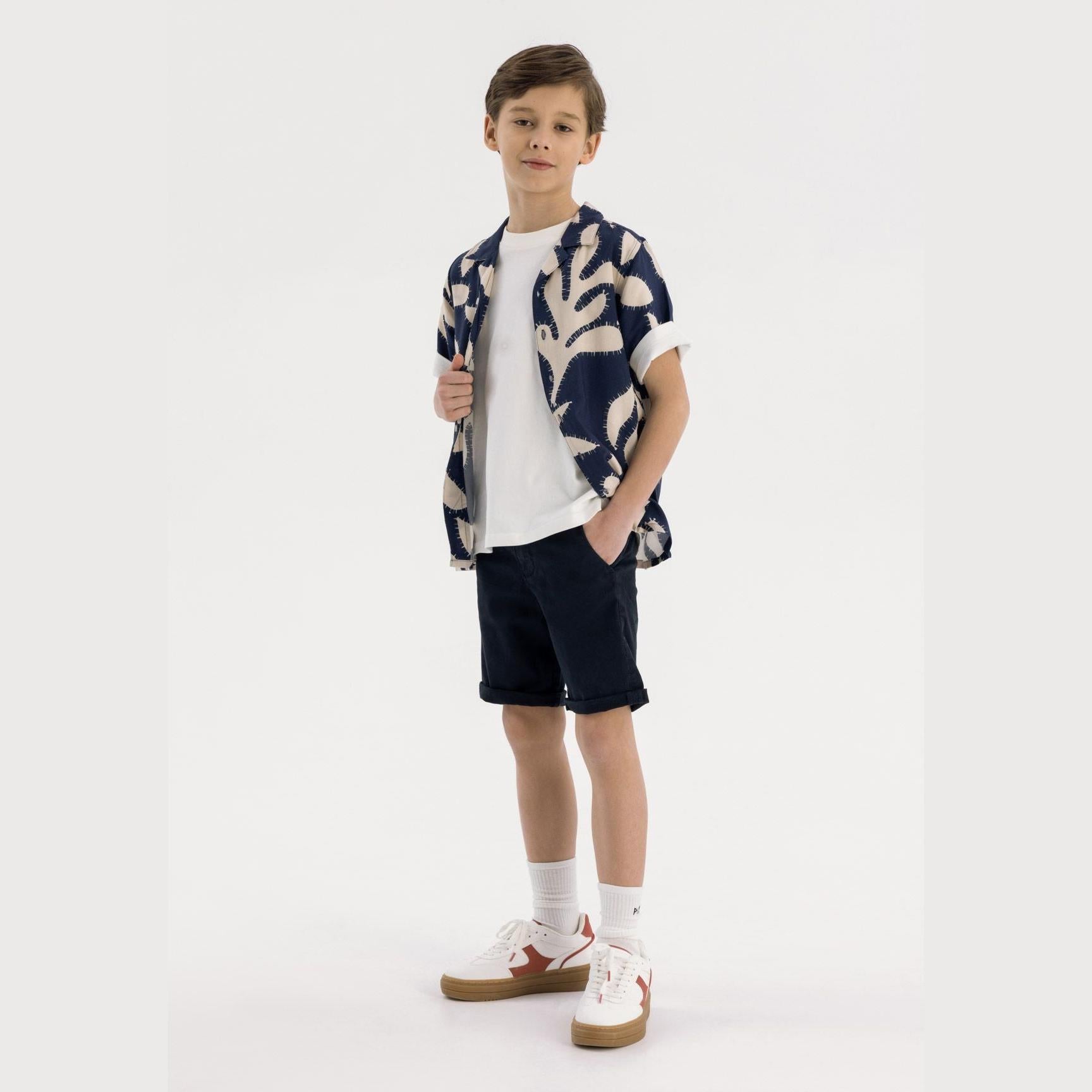 Boy wearing navy chino shorts with printed shirt against white background, full body pose showing casual styling