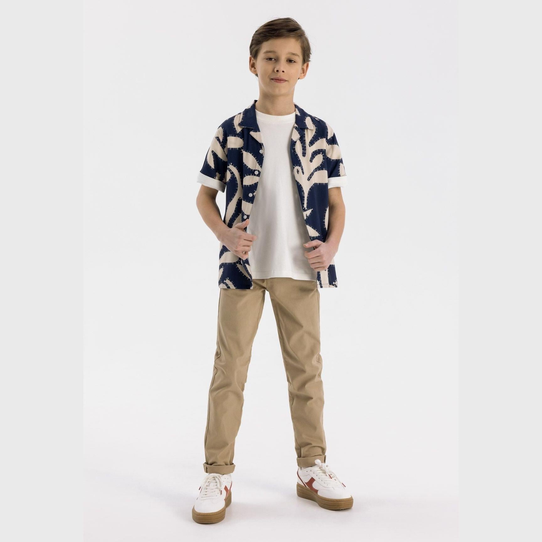 Boy wearing beige chino trousers with navy patterned shirt and white trainers on white background