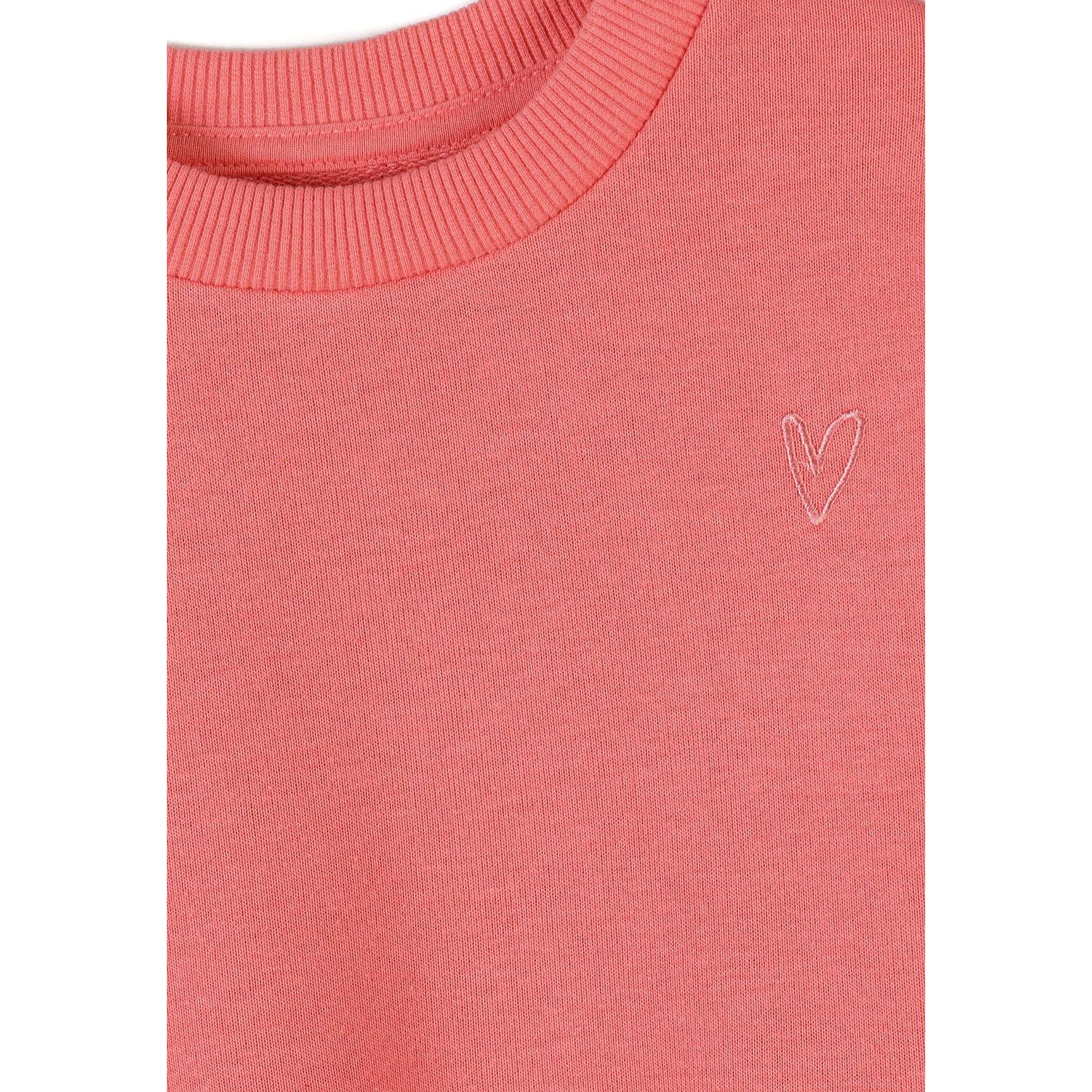 Close-up detail of coral pink sweatshirt showing embroidered white heart motif and ribbed crew neckline