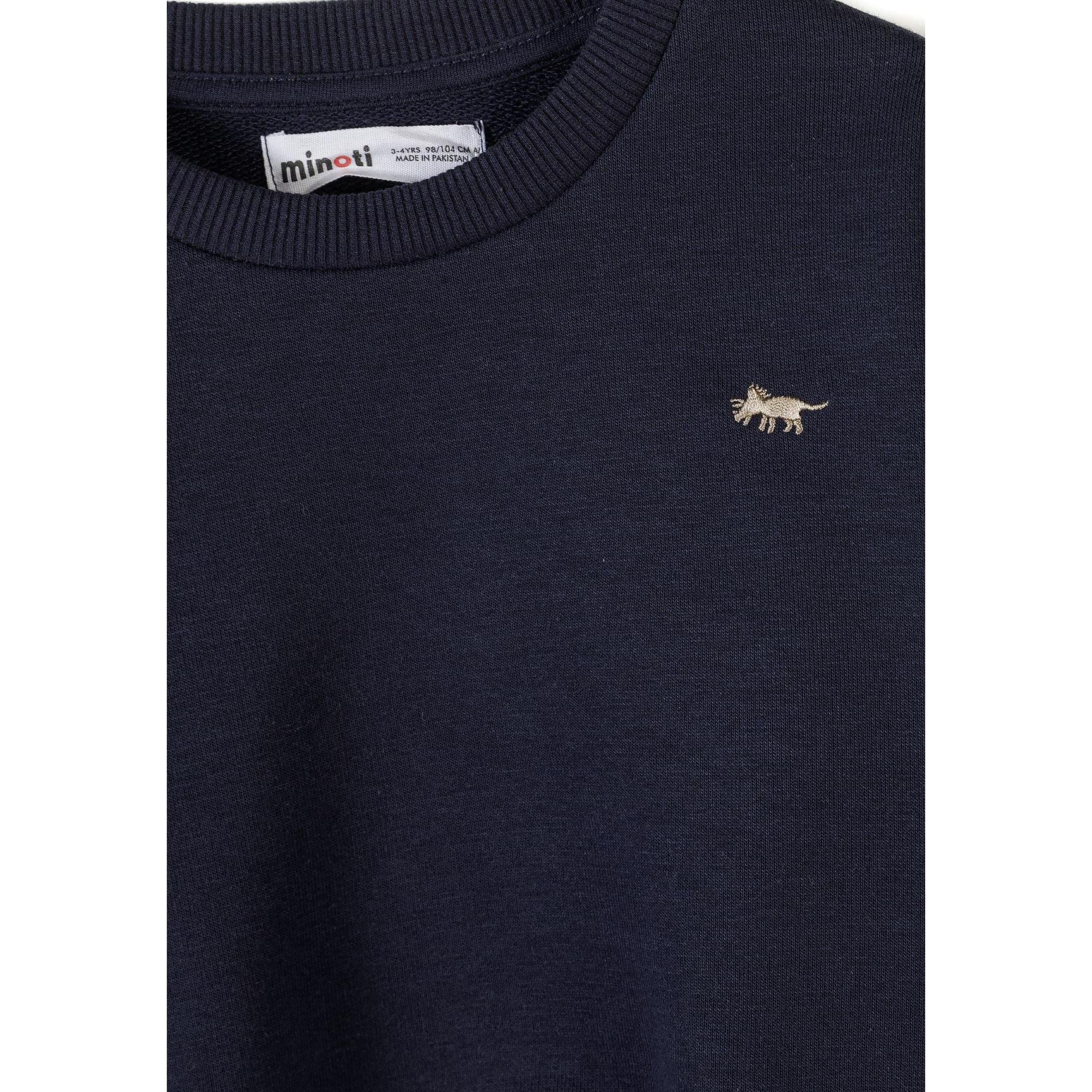 Close-up detail of grey sweatshirt showing Minoti label at neck and small embroidered navy dinosaur logo on chest