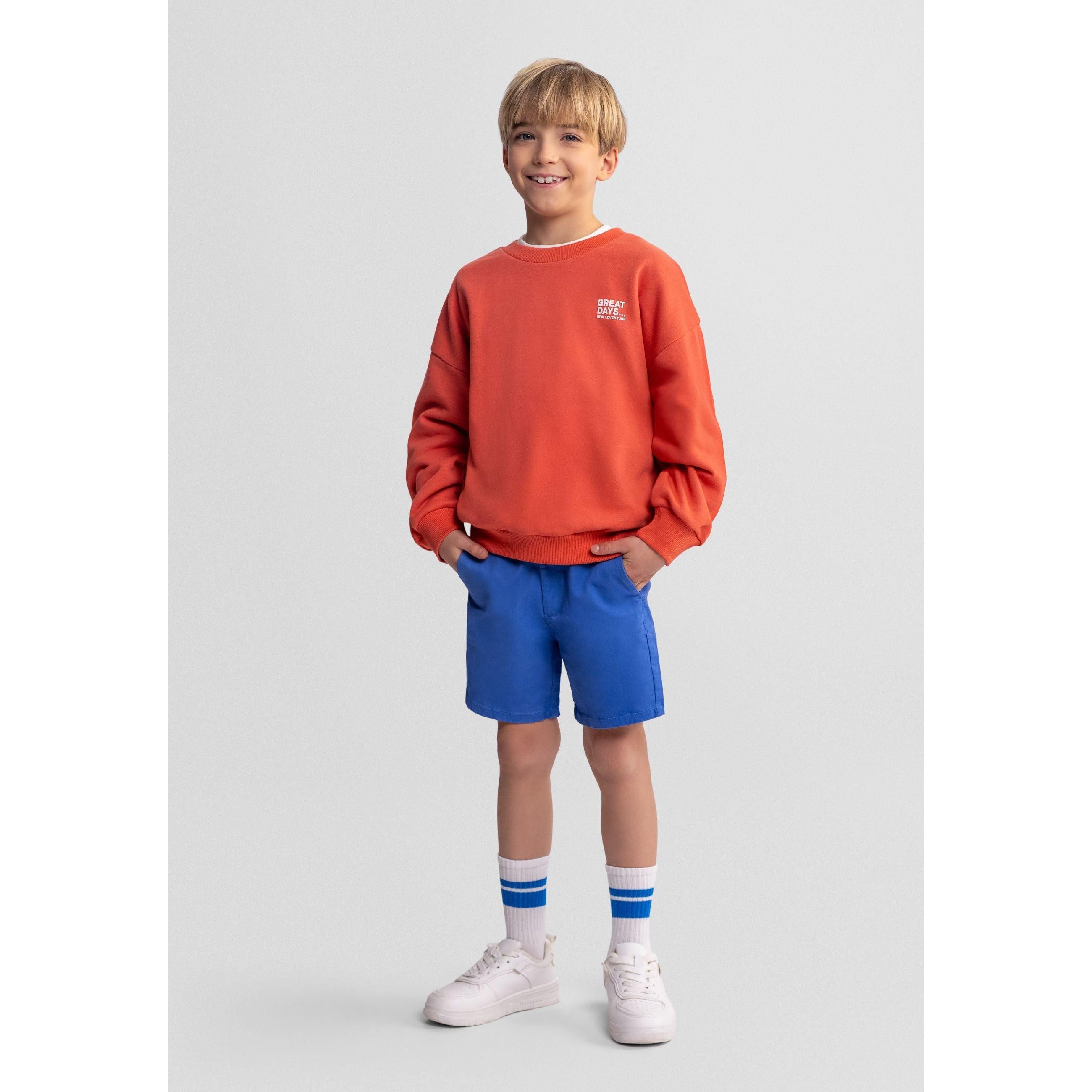 Young blonde boy wearing orange-red sweatshirt with white chest print and blue shorts, standing against white background with hands in pockets and white striped socks