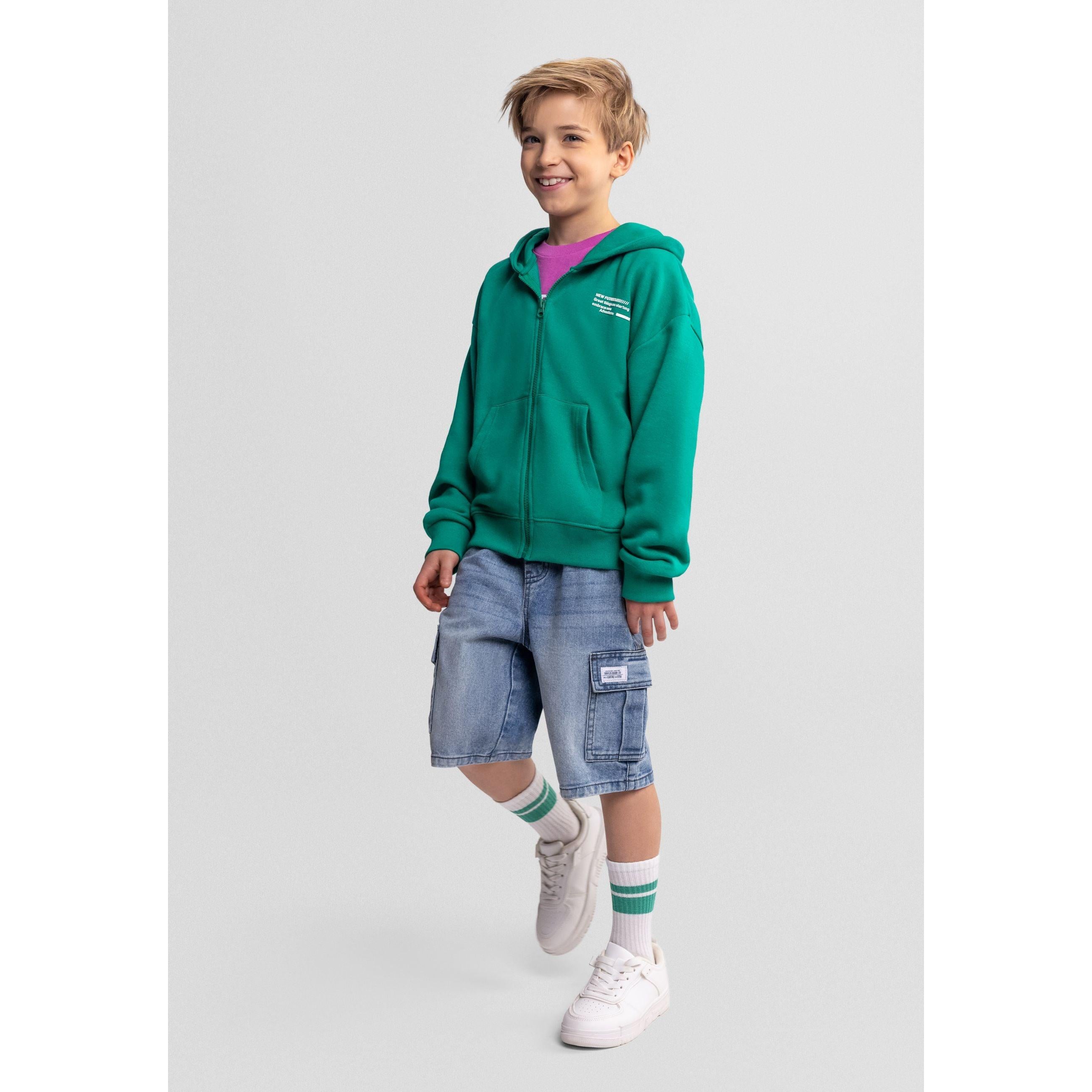 Boy wearing green zip hoodie with denim shorts and striped socks on grey background