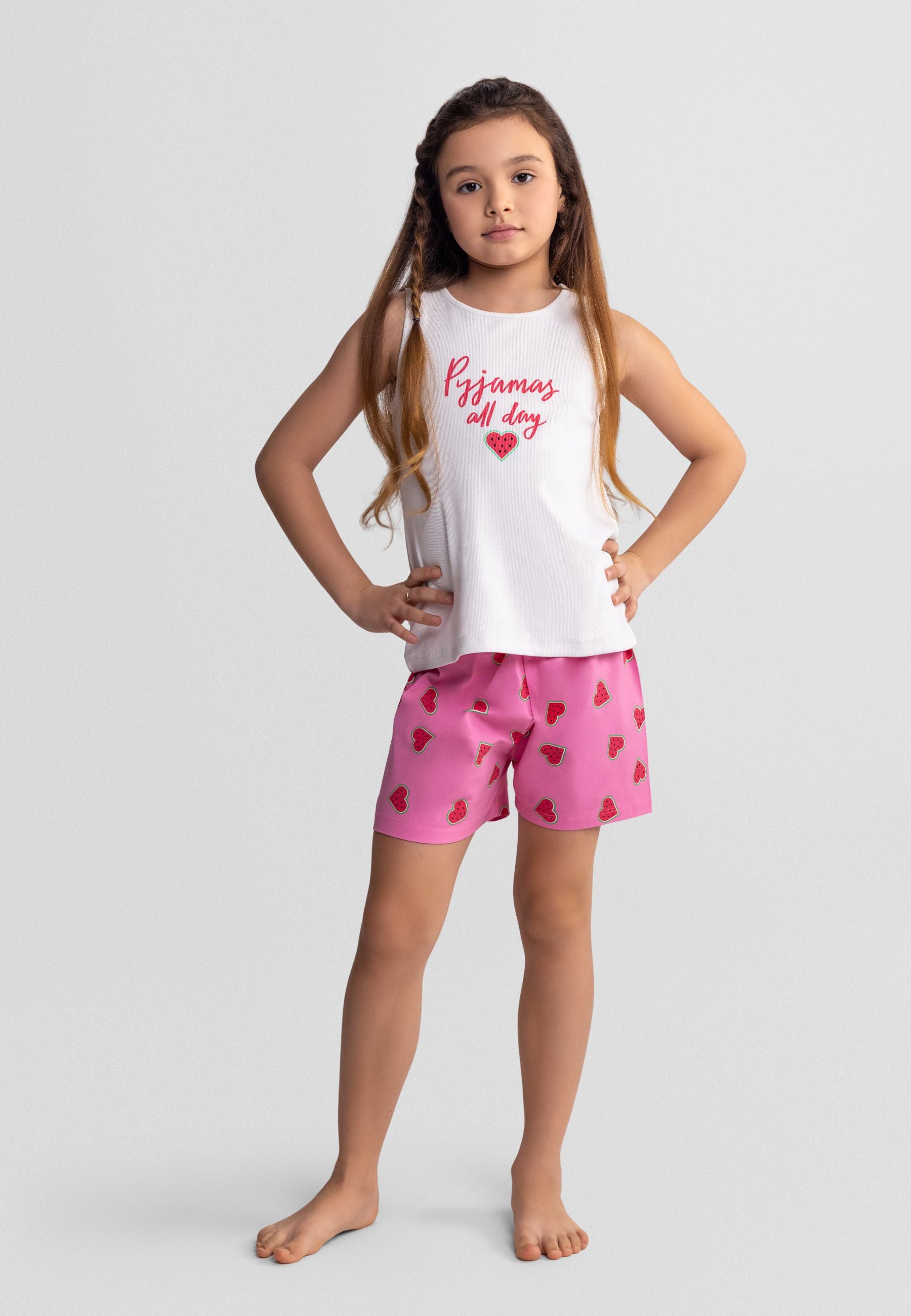 Young girl wearing white ribbed tank top with pink 'Pyjamas all day' text and heart design, paired with pink watermelon print shorts, standing against light background