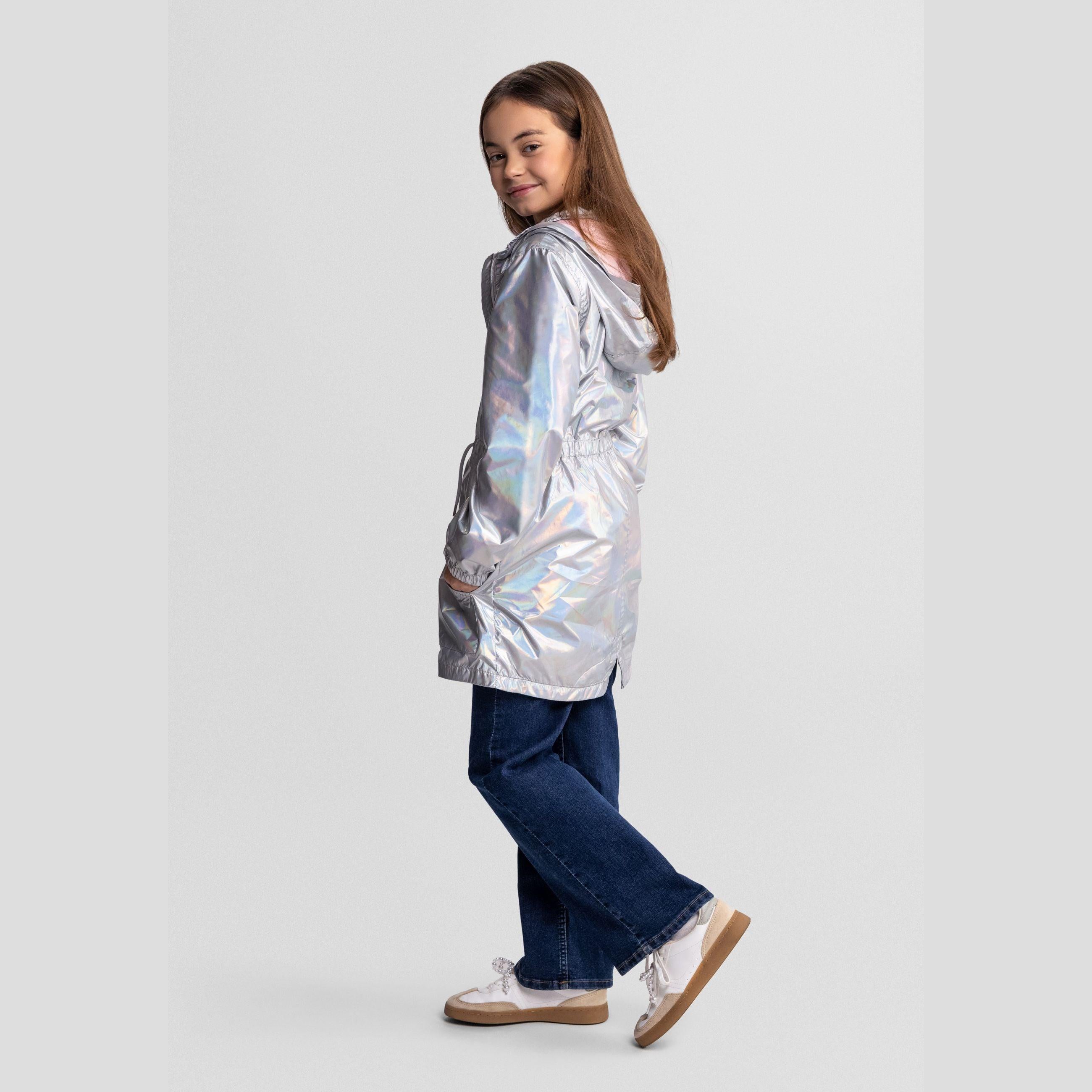 Side profile view of girl in silver iridescent rain jacket showing the relaxed fit and length, paired with dark blue jeans on light background