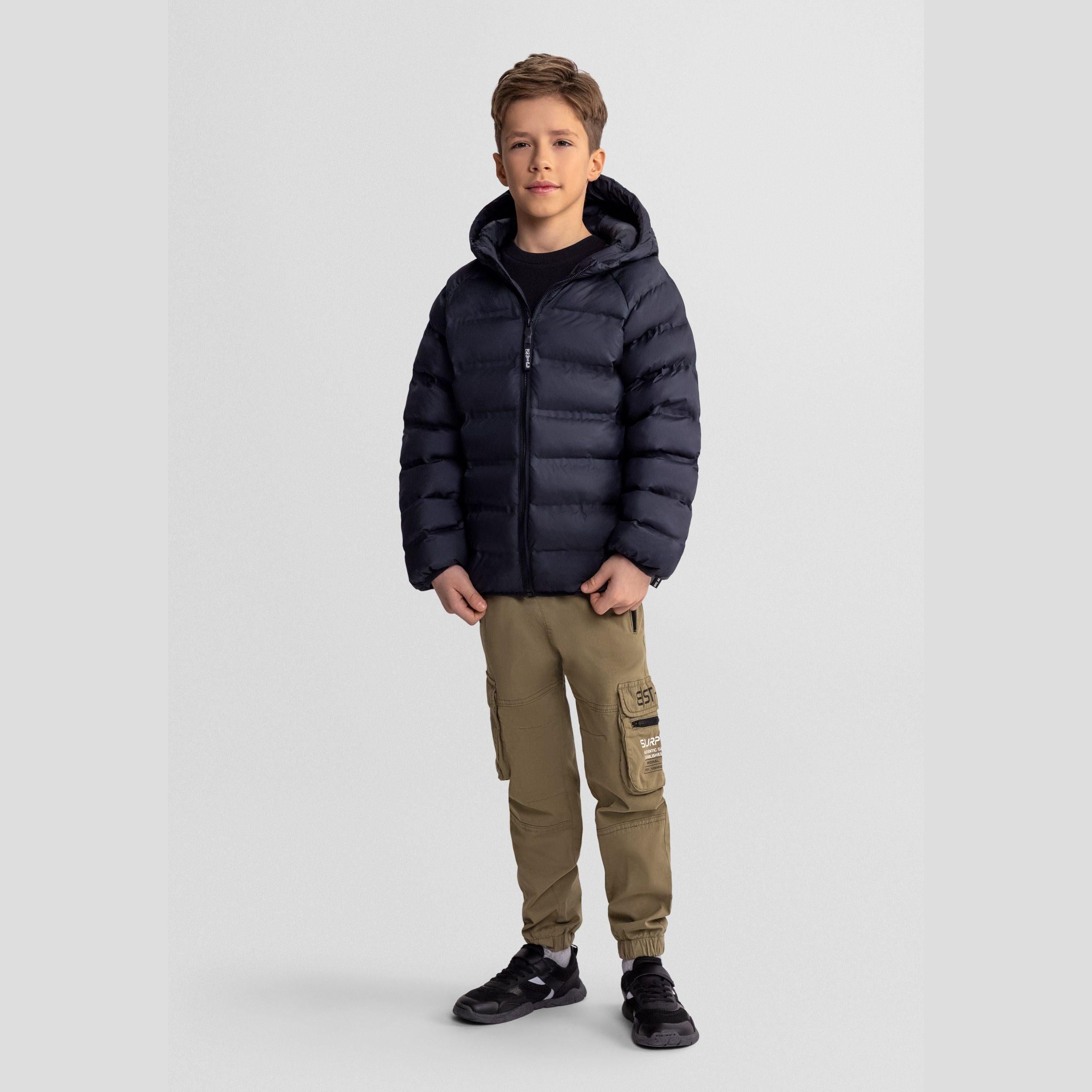 Boy wearing black lightweight padded jacket with hood and khaki cargo pants, standing against light grey studio background, front view full body shot