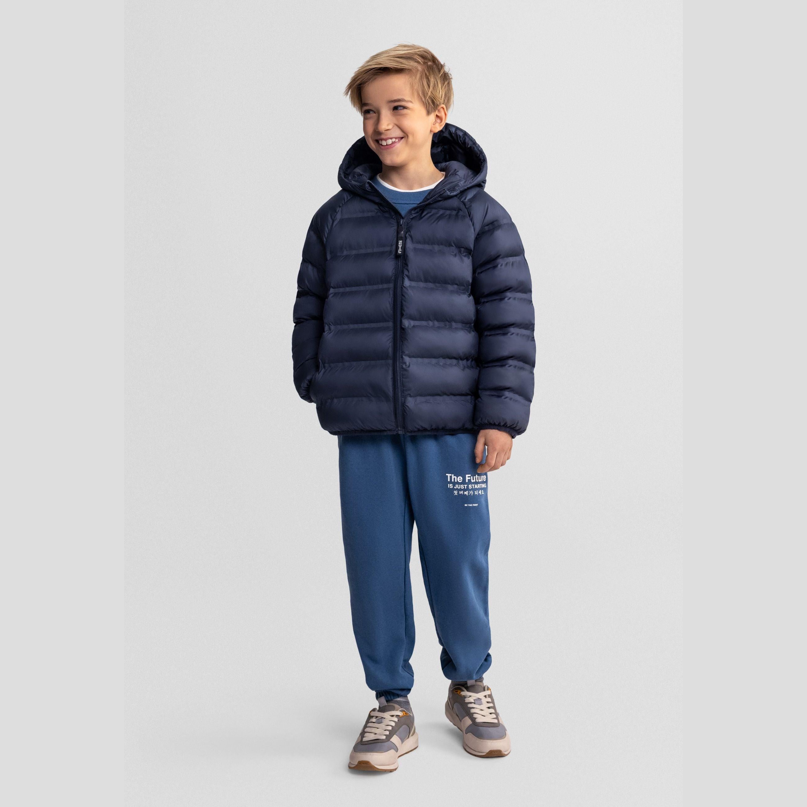 Boy wearing navy blue quilted jacket with hood and blue joggers against light grey background