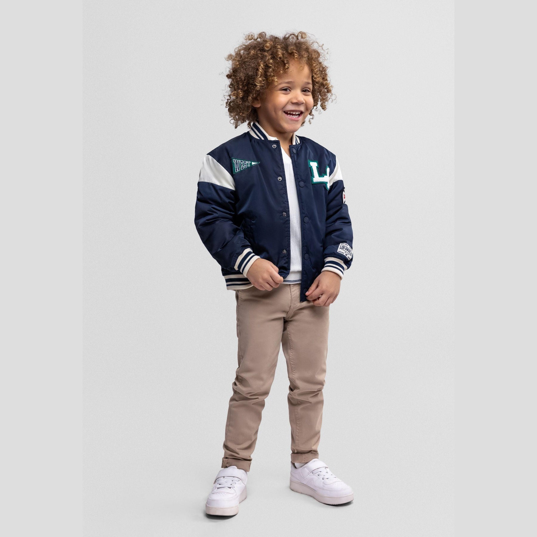 Young child with curly hair wearing navy varsity bomber jacket with white sleeves and beige trousers, smiling against white background
