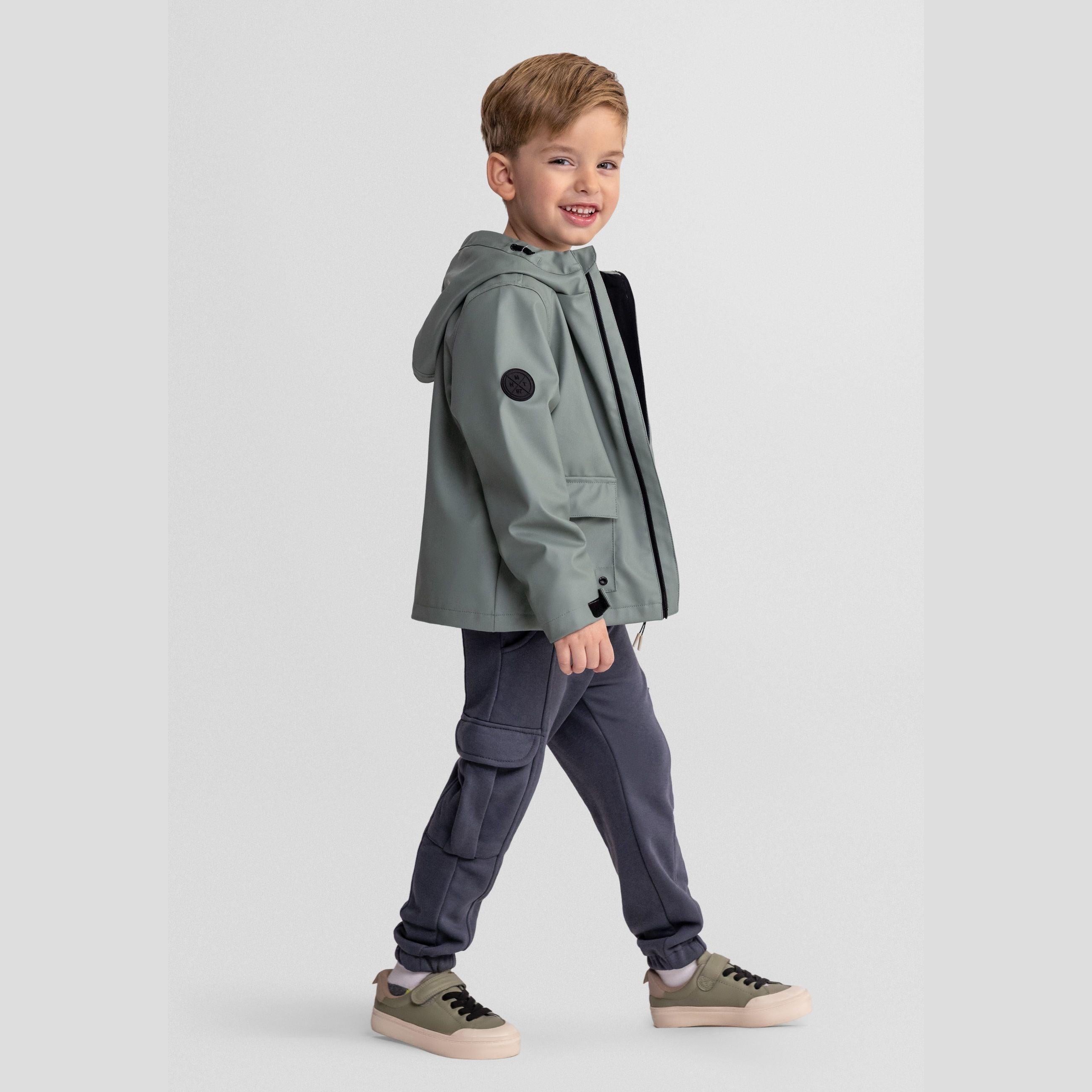 Young boy wearing sage green waterproof parka jacket with hood and navy cargo trousers, walking pose on white background