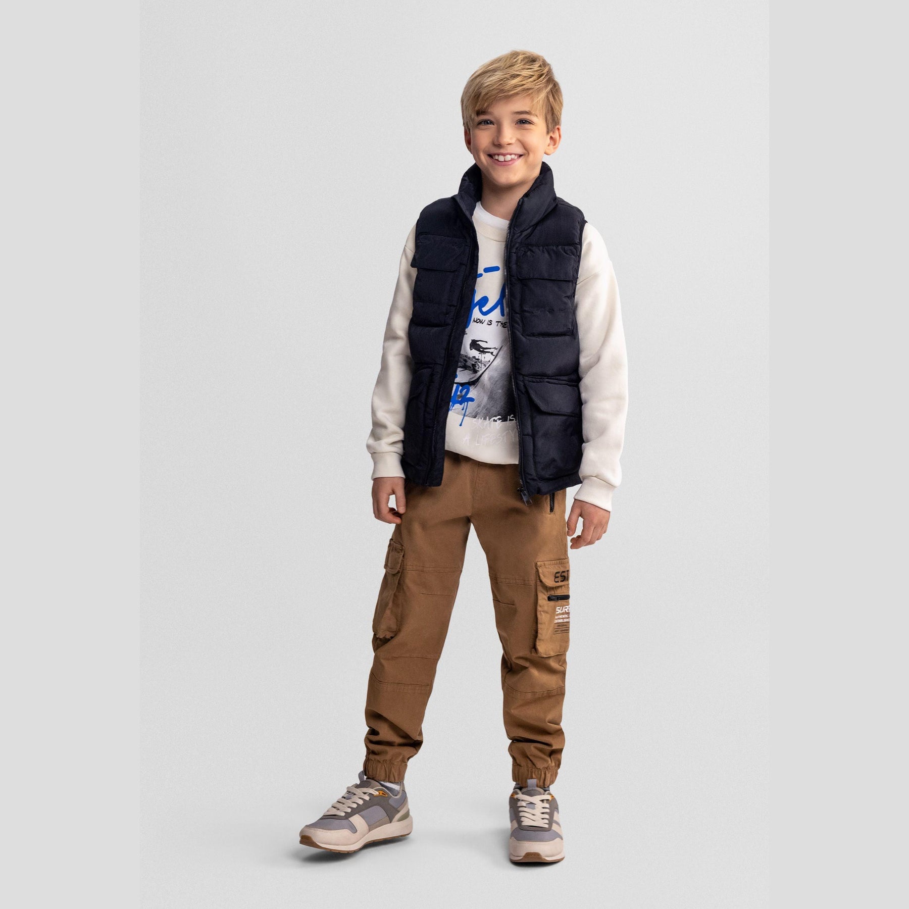 Young boy wearing black quilted waistcoat over white graphic sweatshirt and brown cargo pants, standing against light grey background