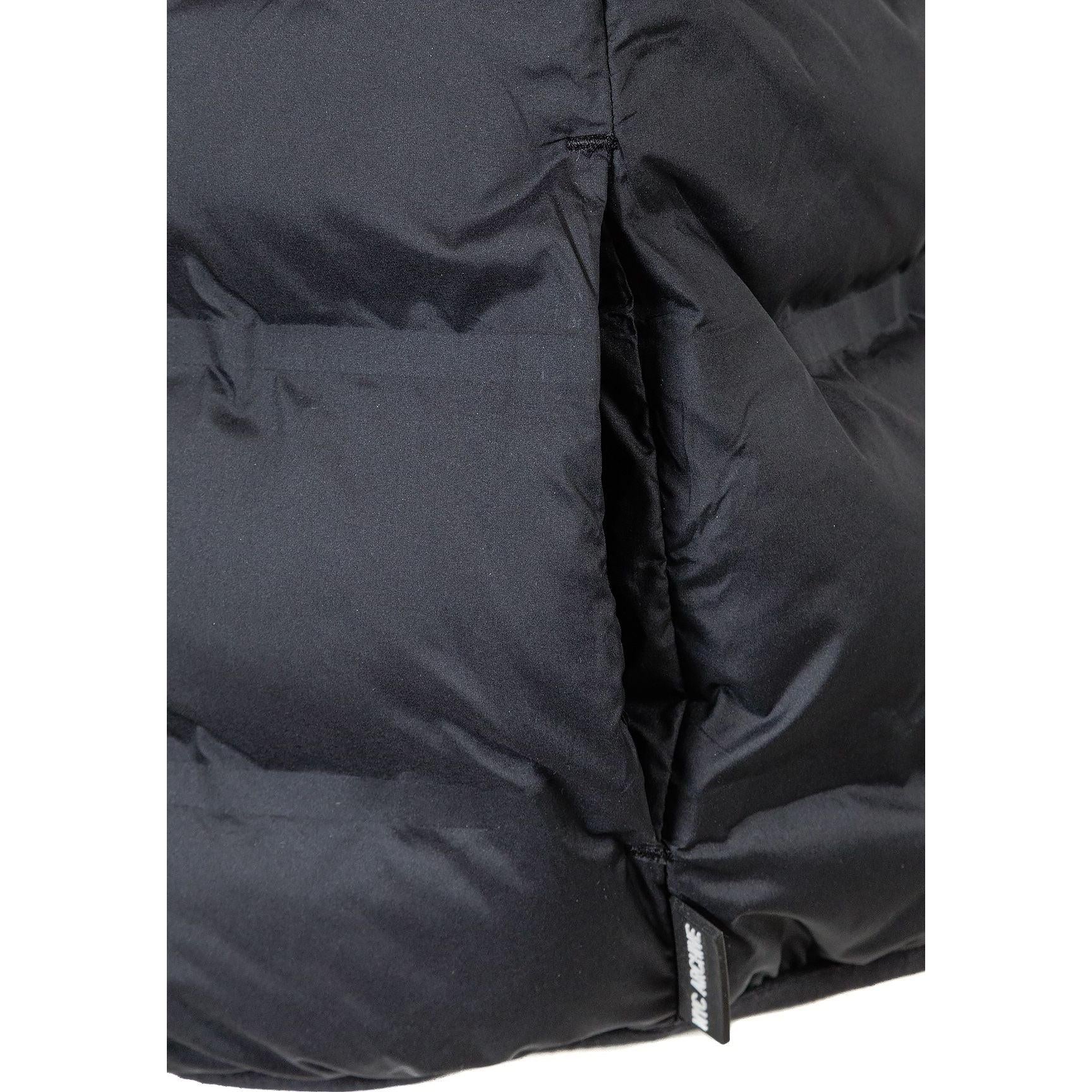 Boy in dynamic pose wearing black quilted waistcoat over beige tracksuit, hands pulling vest open, against light grey studio background