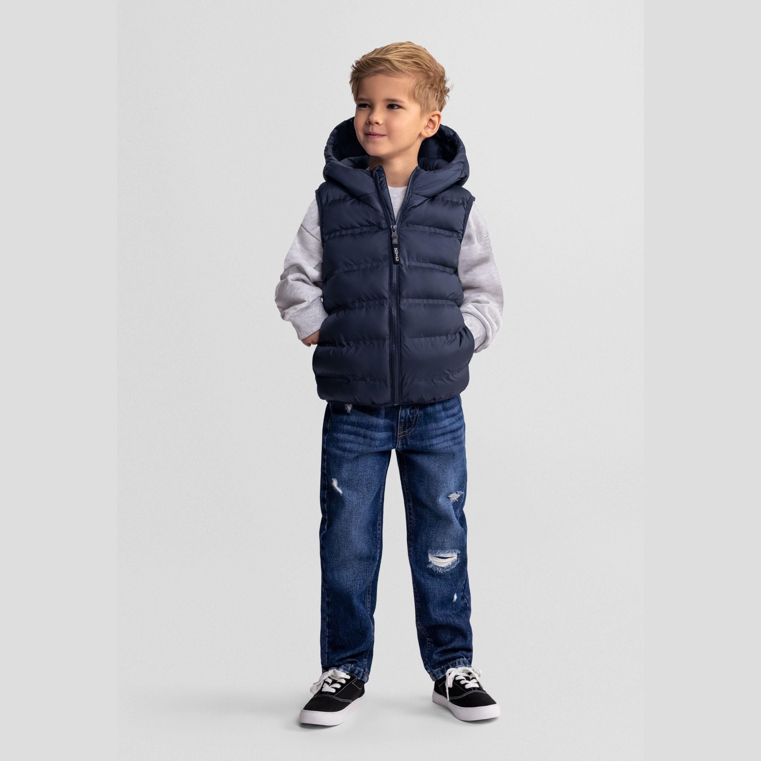 Young boy wearing navy blue hooded padded gilet over light grey hoodie with distressed blue jeans and black sneakers, standing against light grey background