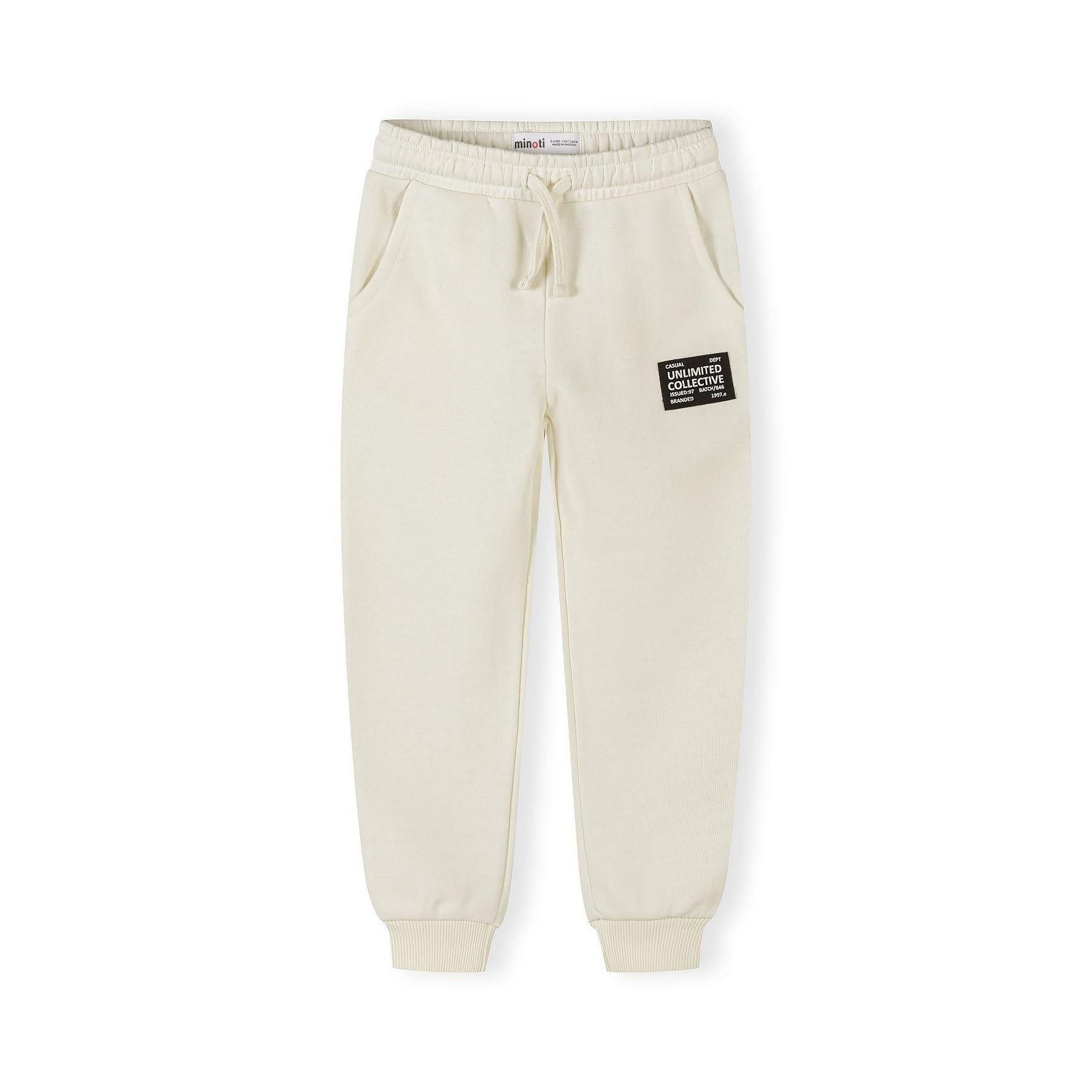 Close-up of cream joggers with black branded patch on thigh, elastic waistband with drawstring, and ribbed cuffs on white background