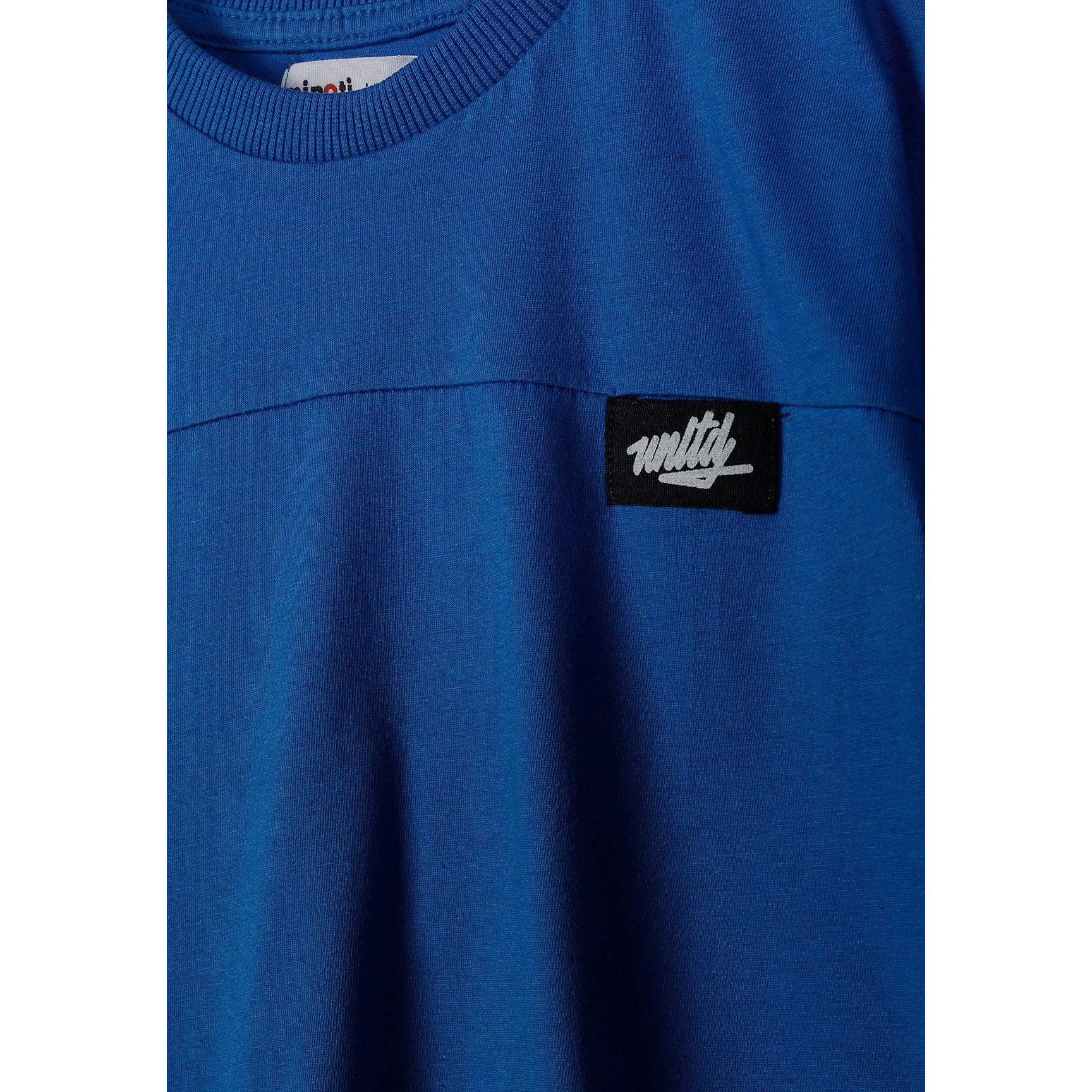 Close-up detail of royal blue t-shirt showing ribbed crew neckline and black rectangular Minoti logo patch with white text