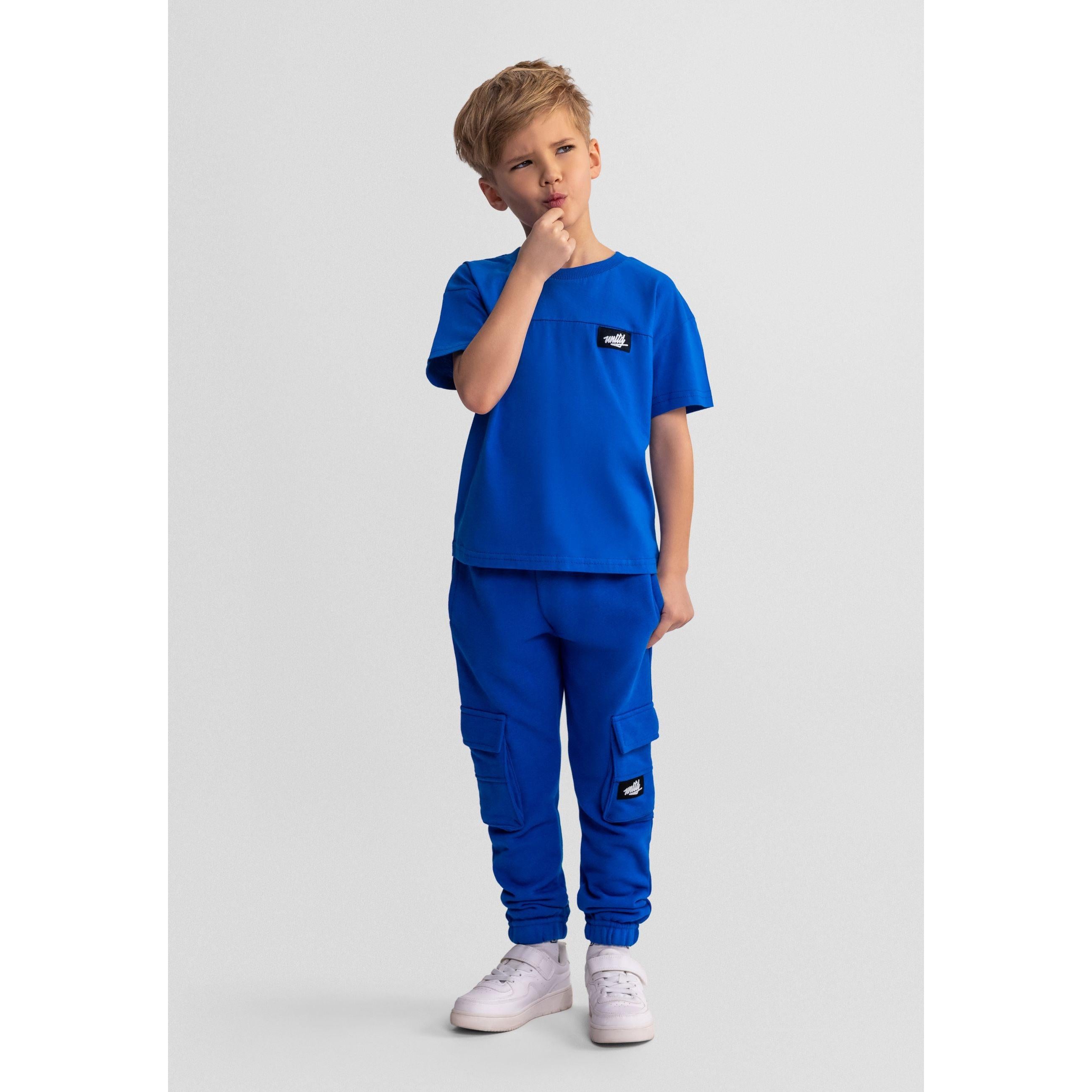 Boy wearing royal blue jogging set with t-shirt and cargo joggers, standing against grey background with hand to chin in thinking pose