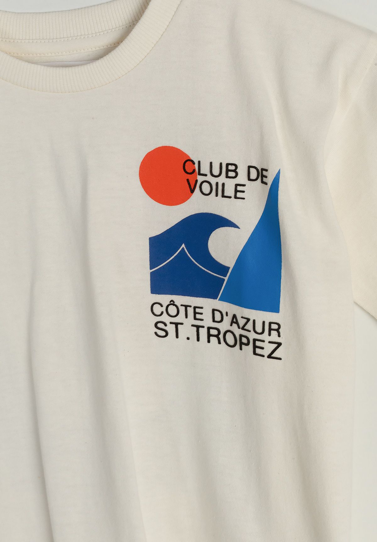 Close-up of Club de Voile chest print with orange sun, blue sail and waves on cream T-shirt.