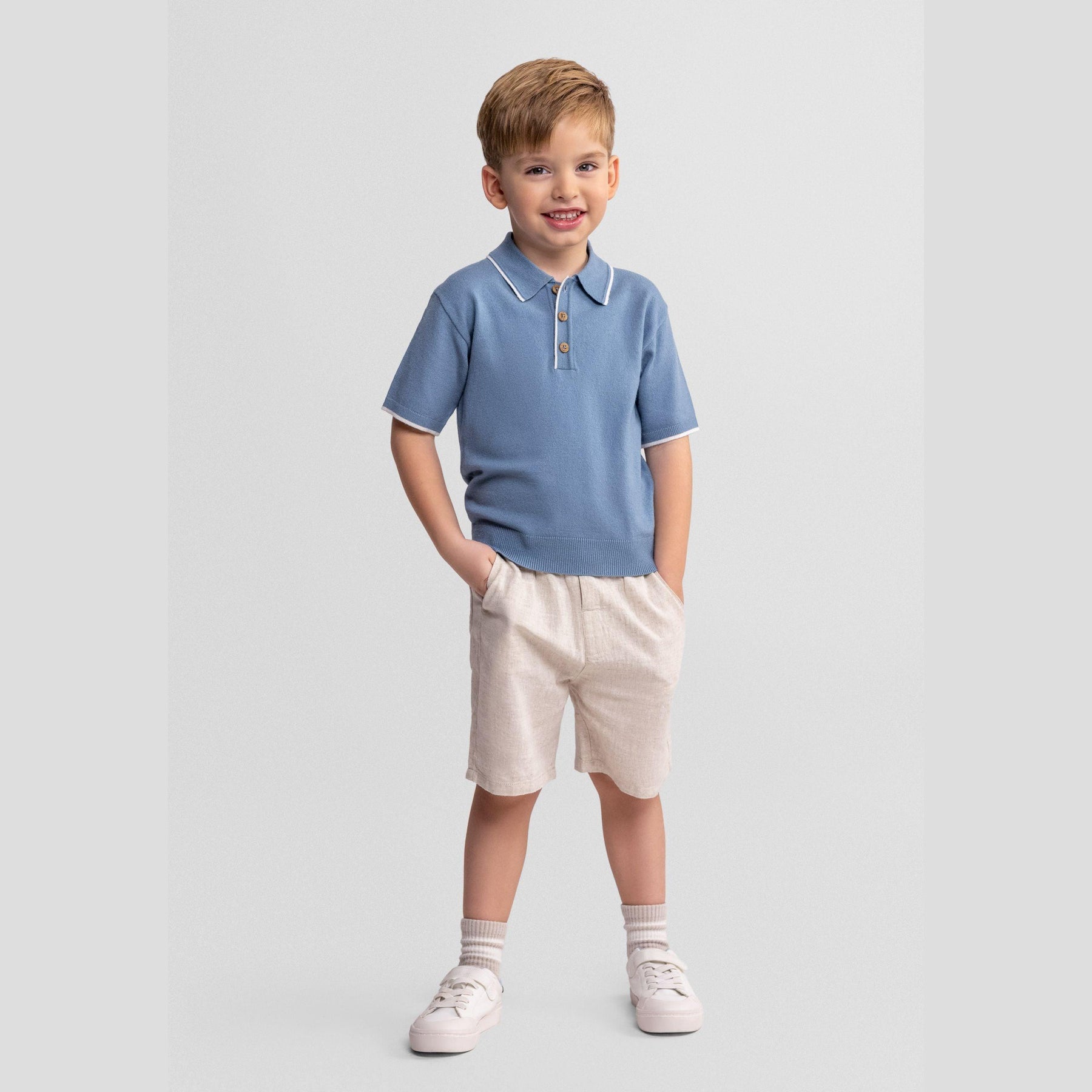 Young boy wearing light blue knitted polo shirt and beige linen shorts, standing against white background with hands in pockets, smiling at camera