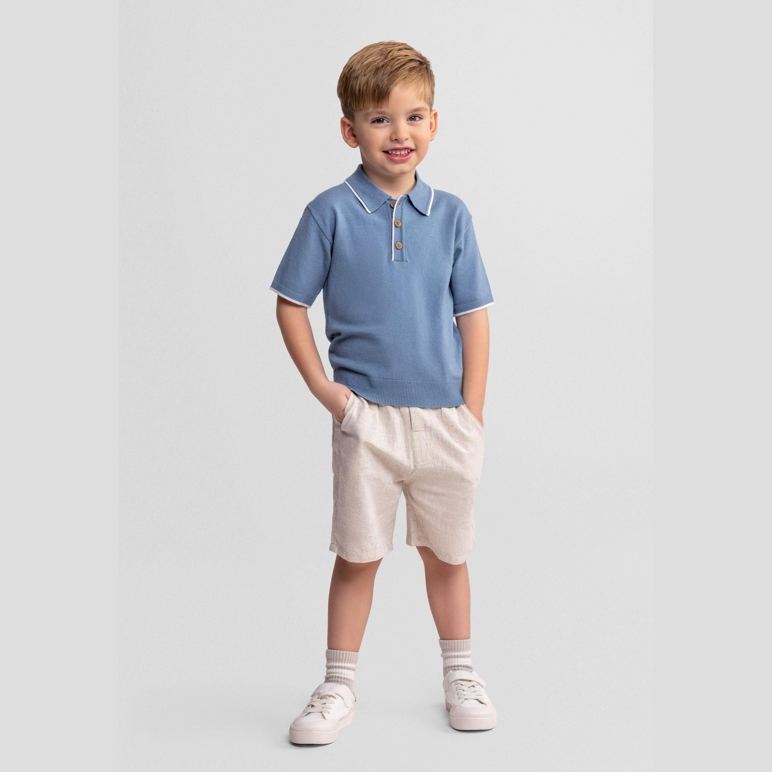 Young boy wearing light blue knitted polo shirt and beige linen shorts, standing against white background with hands in pockets, smiling at camera