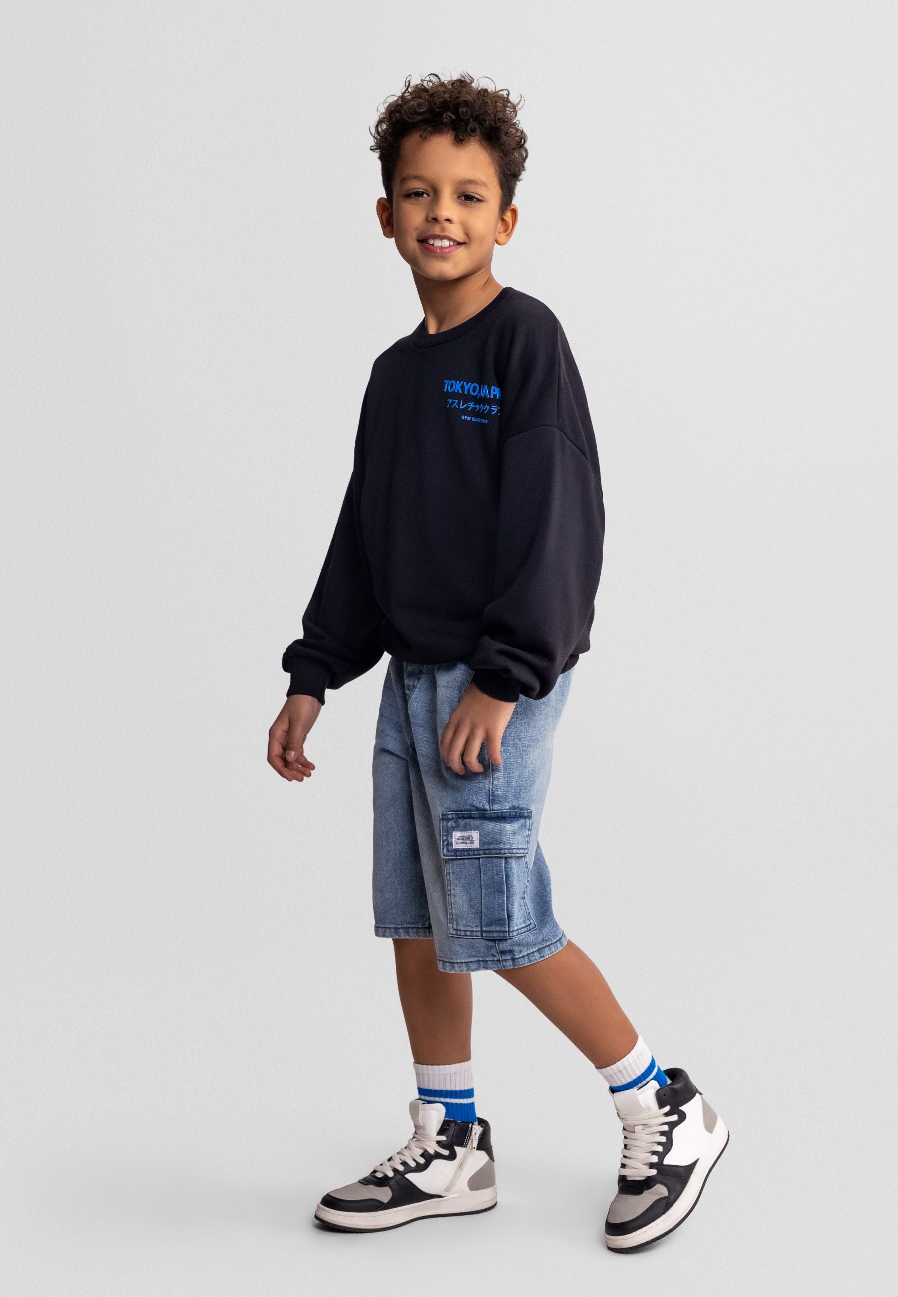 Young boy wearing navy sweatshirt and blue denim cargo shorts, standing against grey background with hands at sides