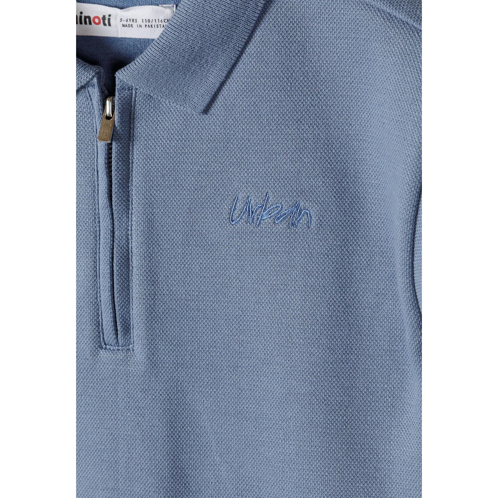 Close-up detail of blue polo shirt showing zip collar, embroidered Minoti logo, and pique texture with brand label visible