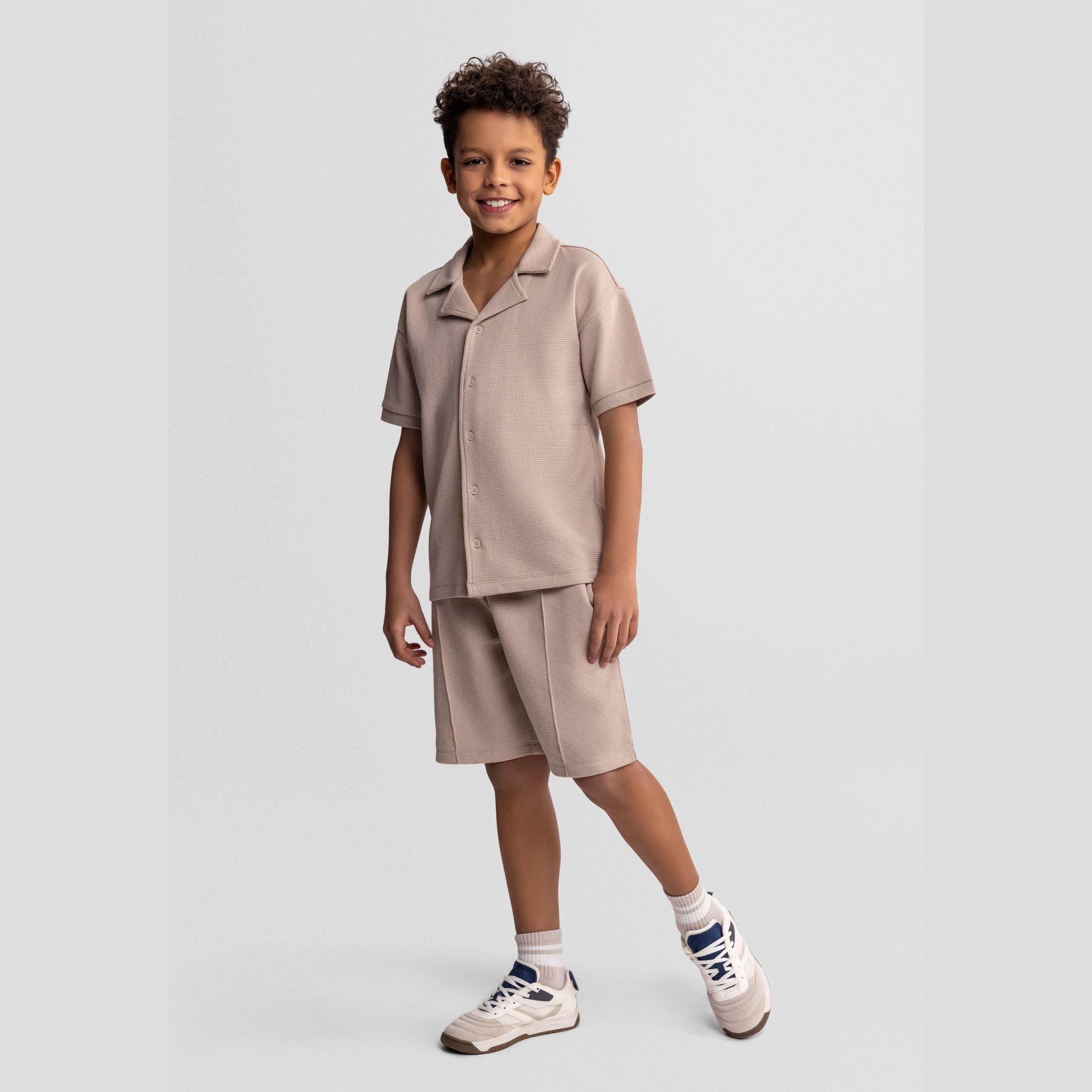 Young boy wearing taupe waffle cotton shirt and shorts set, standing against light grey background, smiling at camera