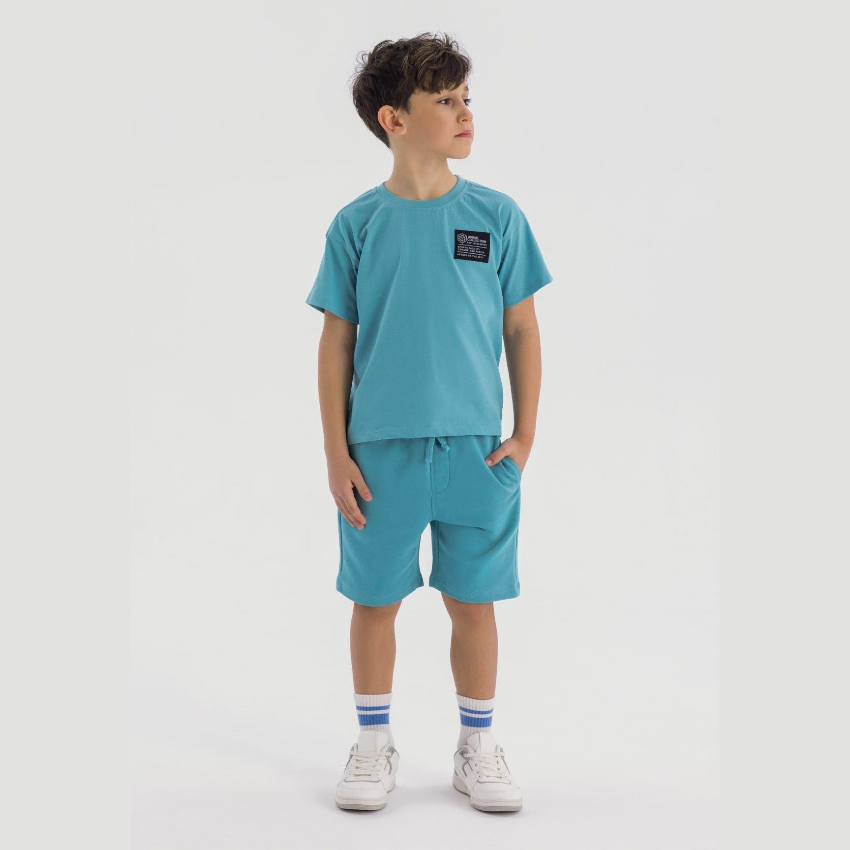 Boy wearing teal t-shirt and shorts set with white sneakers and striped socks, standing against white background