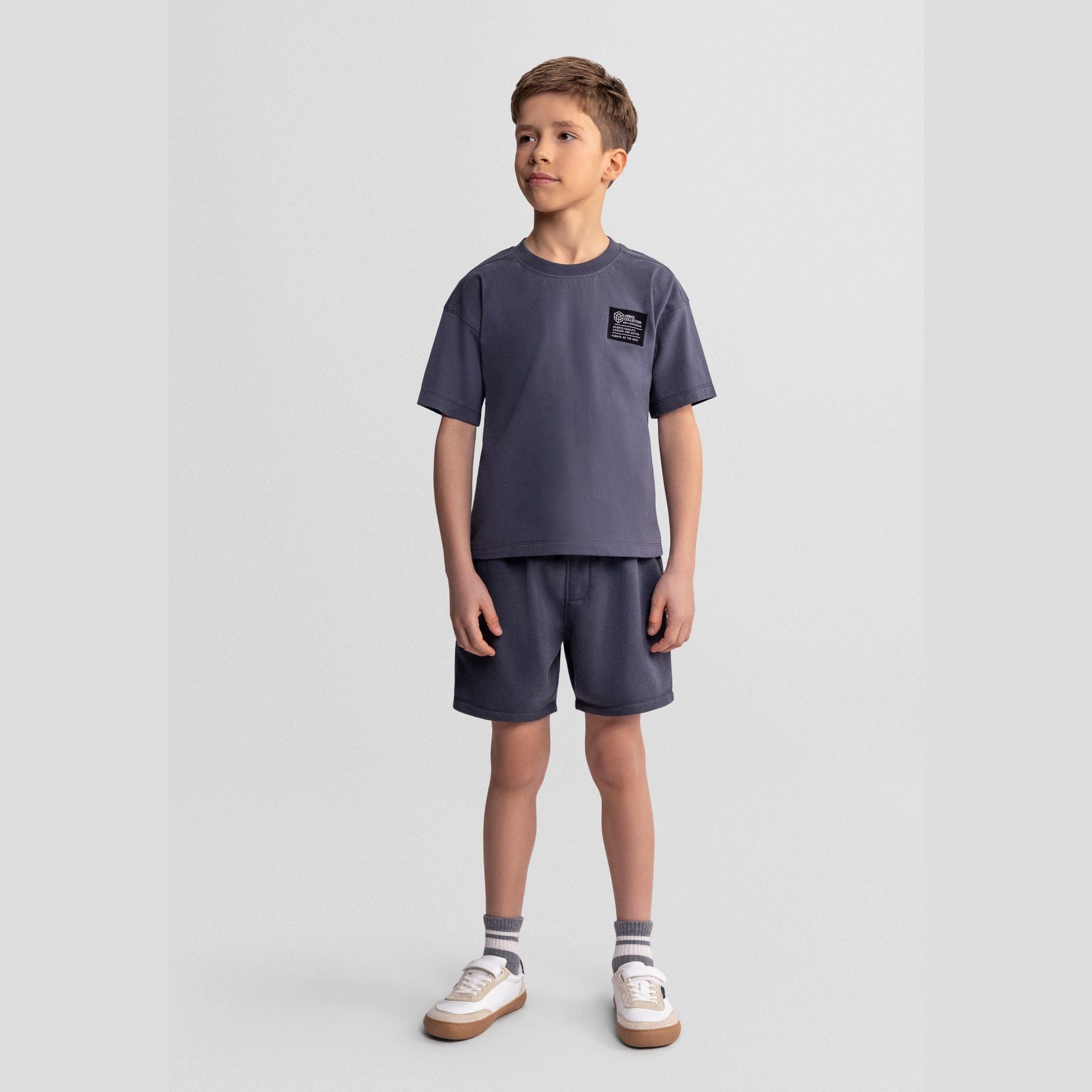 Boy wearing grey cotton t-shirt and matching shorts set with urban patch detail, standing against light background