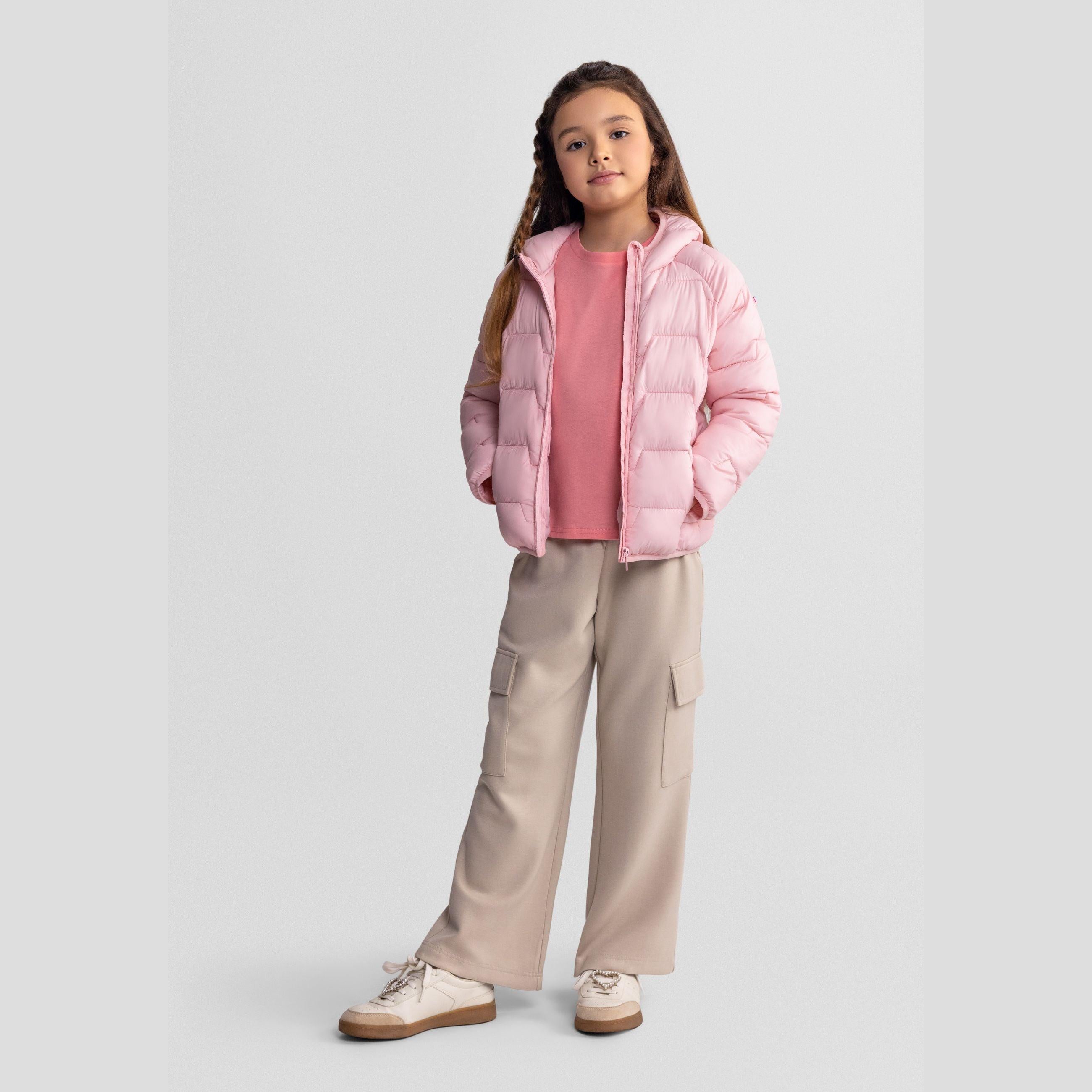 Young girl wearing pink padded hooded jacket over coral top with beige cargo trousers and white sneakers, standing against light grey background