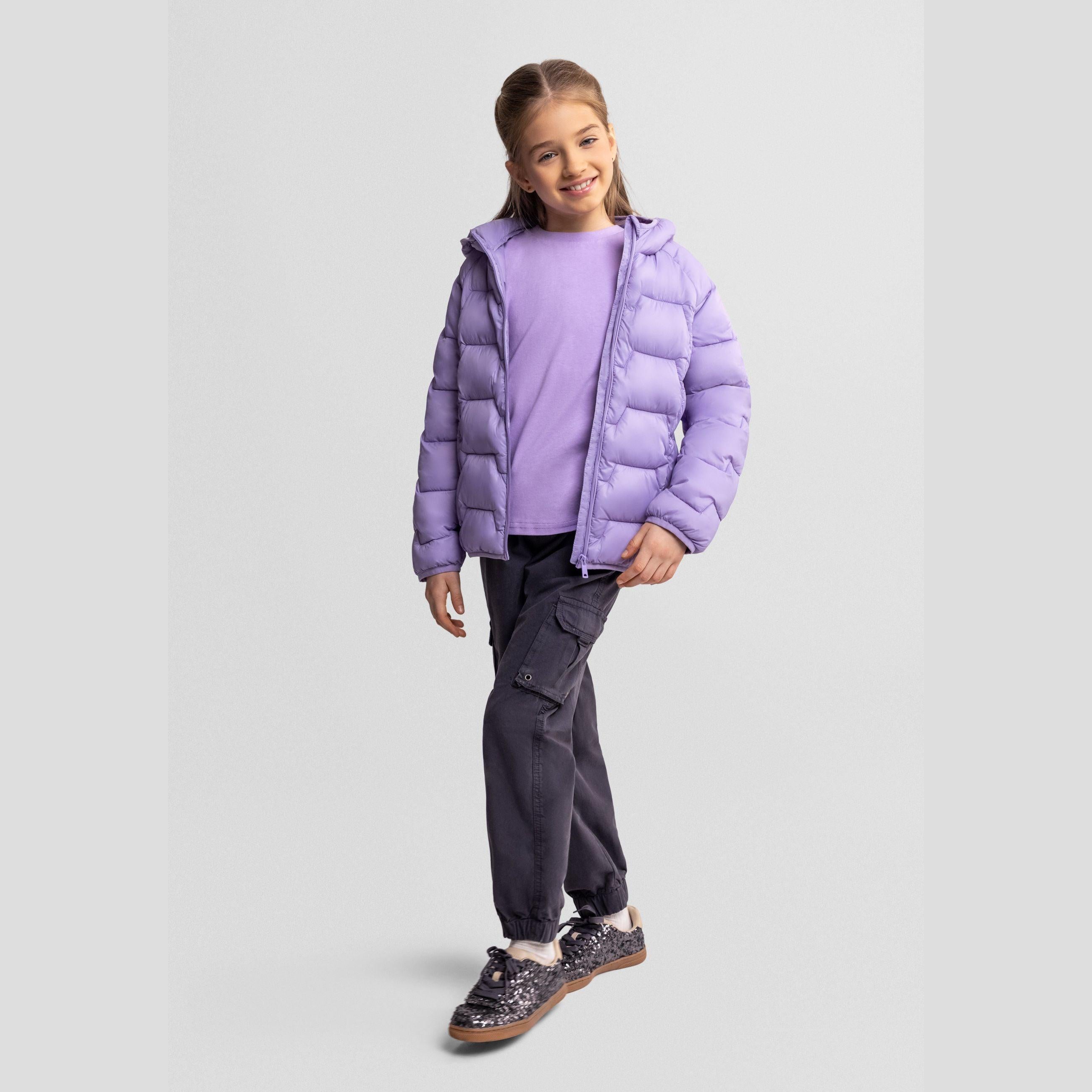 Girl wearing purple lightweight puffer jacket with hood, grey cargo pants and sparkly trainers, smiling against white background